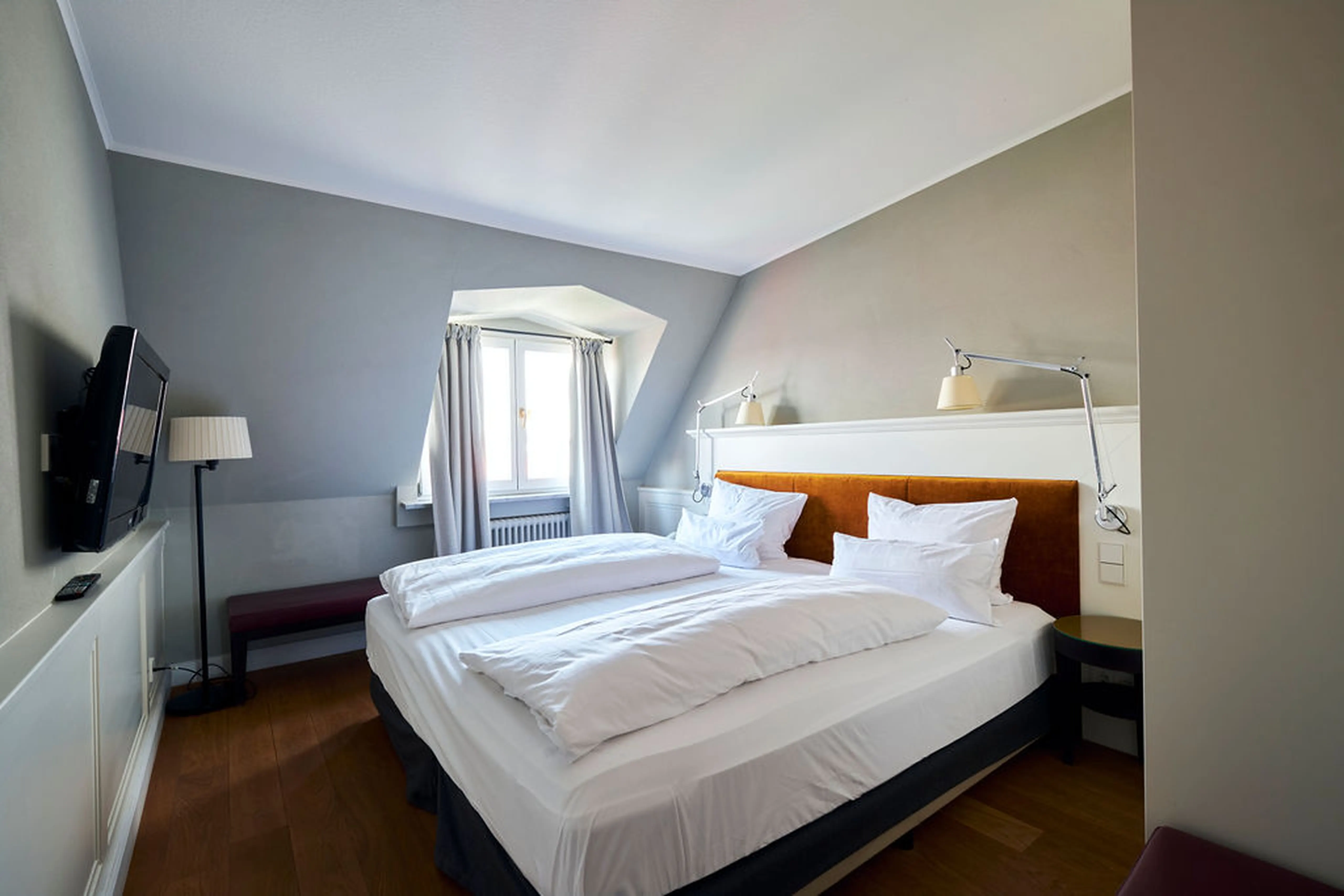 Gemütliches Hotelzimmer mit Doppelbett, zwei Nachttischlampen, einem Fenster und einem Flachbildfernseher an der Wand. Holzboden und dezente Farben.