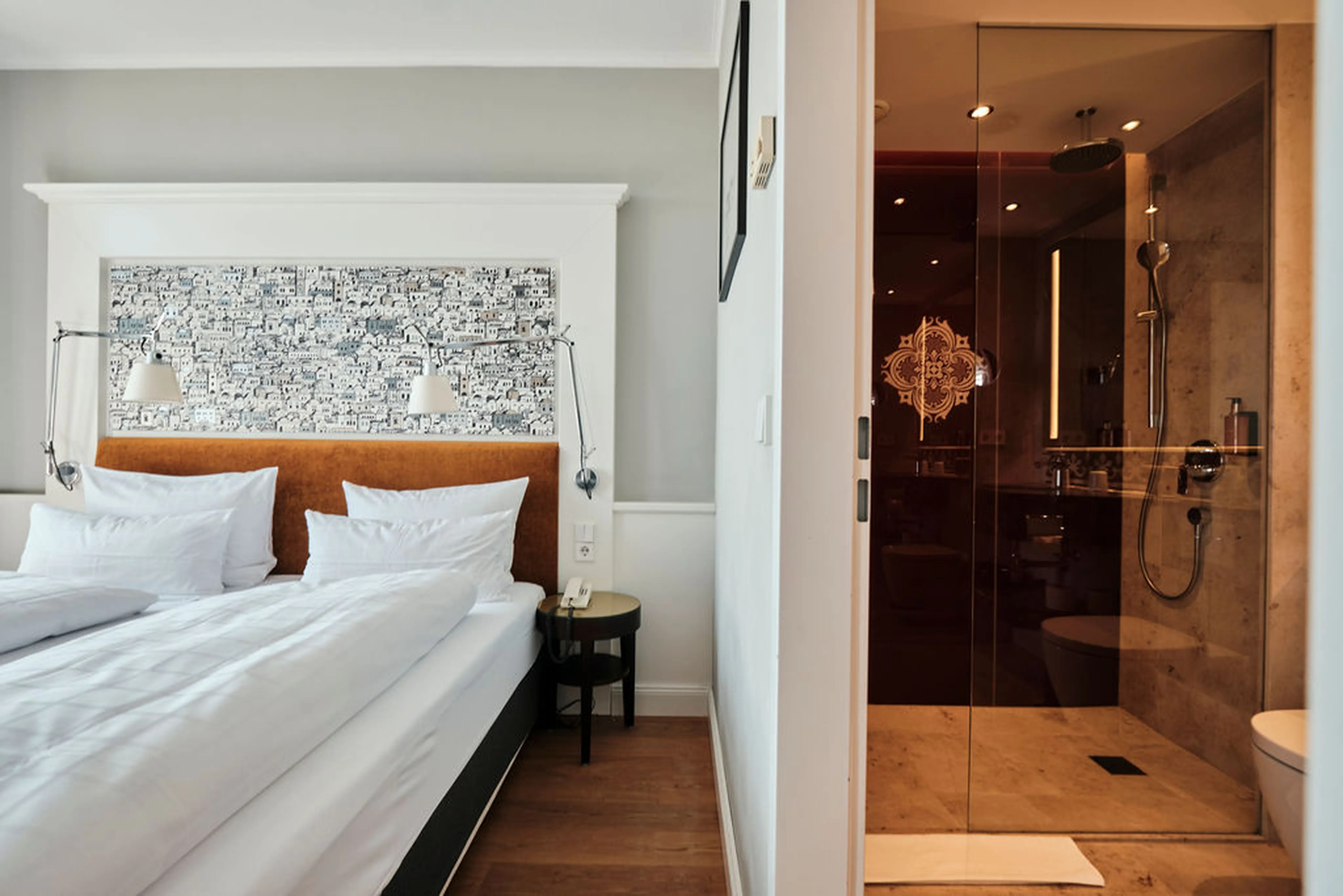 Modernes Hotelzimmer mit Doppelbett, weißer Bettwäsche, dekorativer Tapete, Nachttischlampen und einer angrenzenden Glasdusche im Badezimmer.