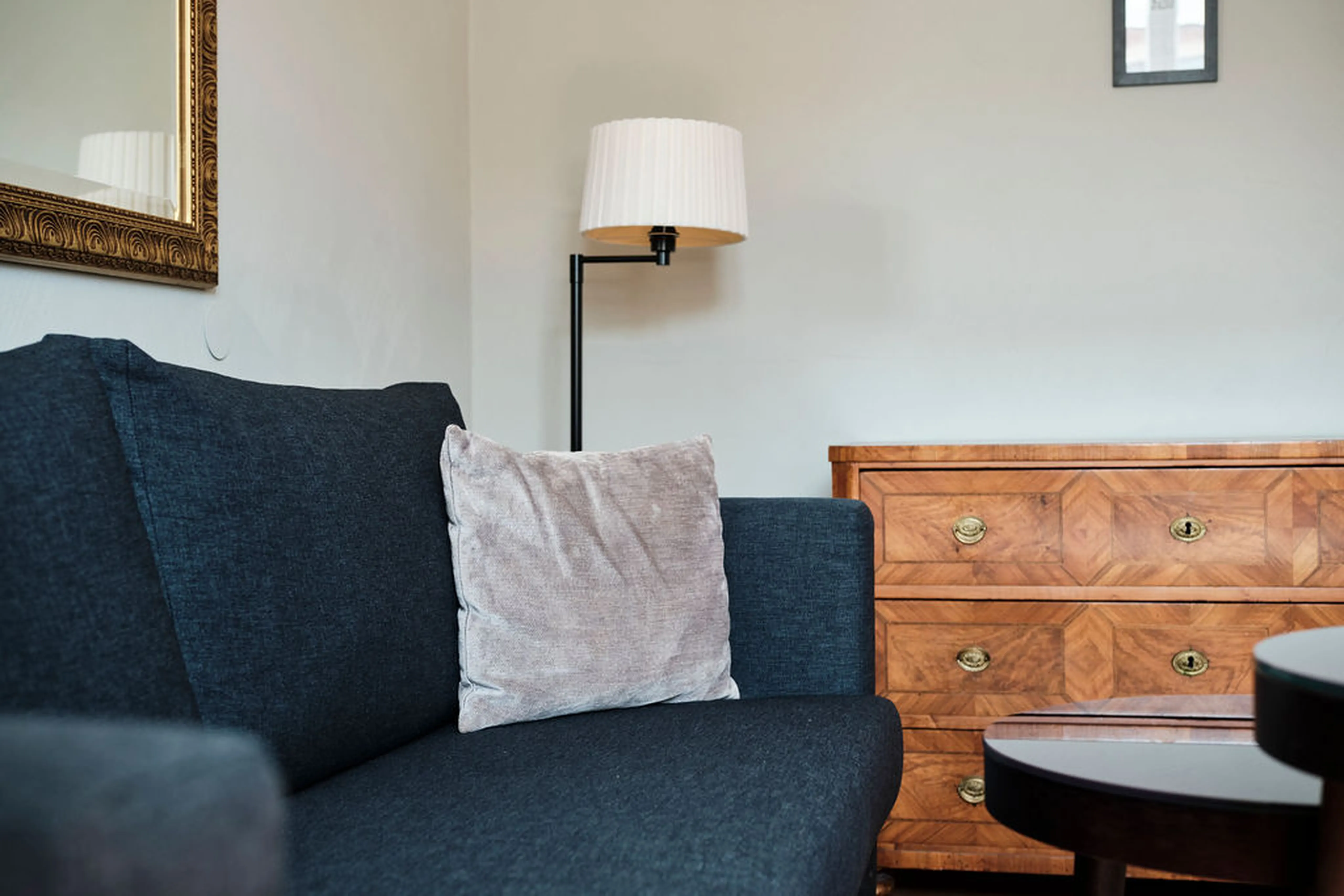 Graues Sofa mit Kissen, Holzkommode und Stehlampe in einem Wohnzimmer. Ein Spiegel hängt an der Wand, ein Tisch ist teilweise sichtbar.