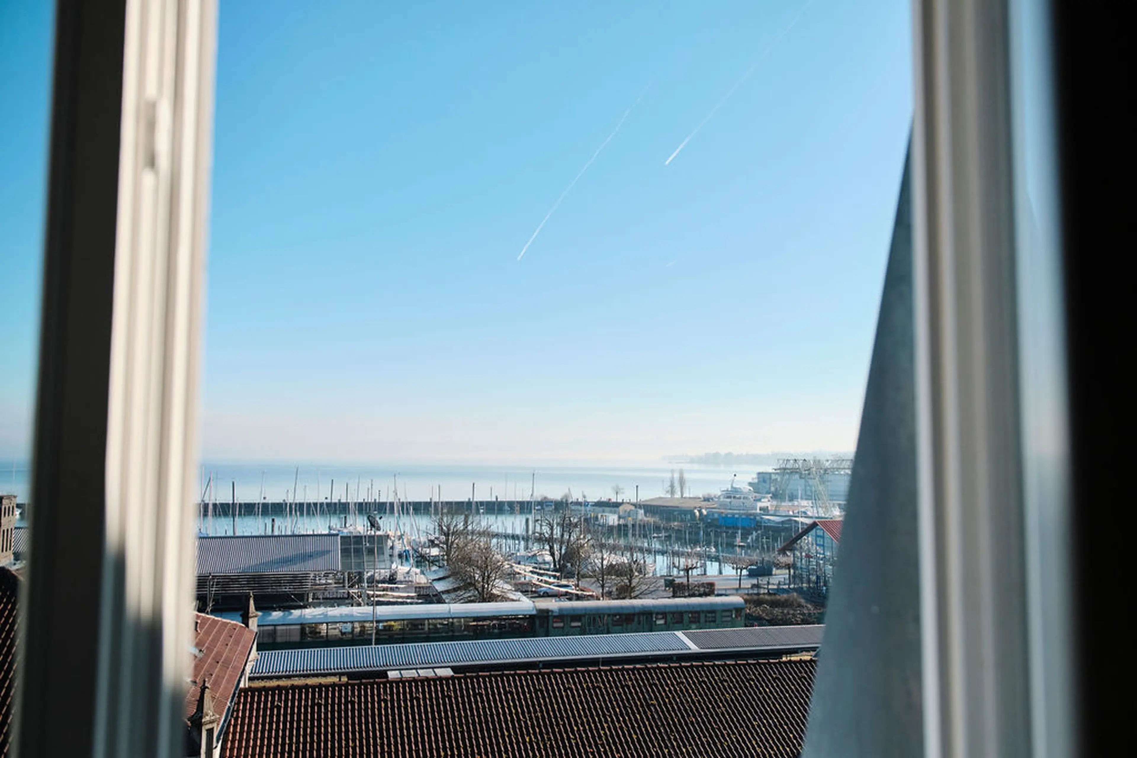 Blick aus einem Fenster auf einen Hafen mit Booten unter blauem Himmel, umgeben von Gebäuden und Bäumen.