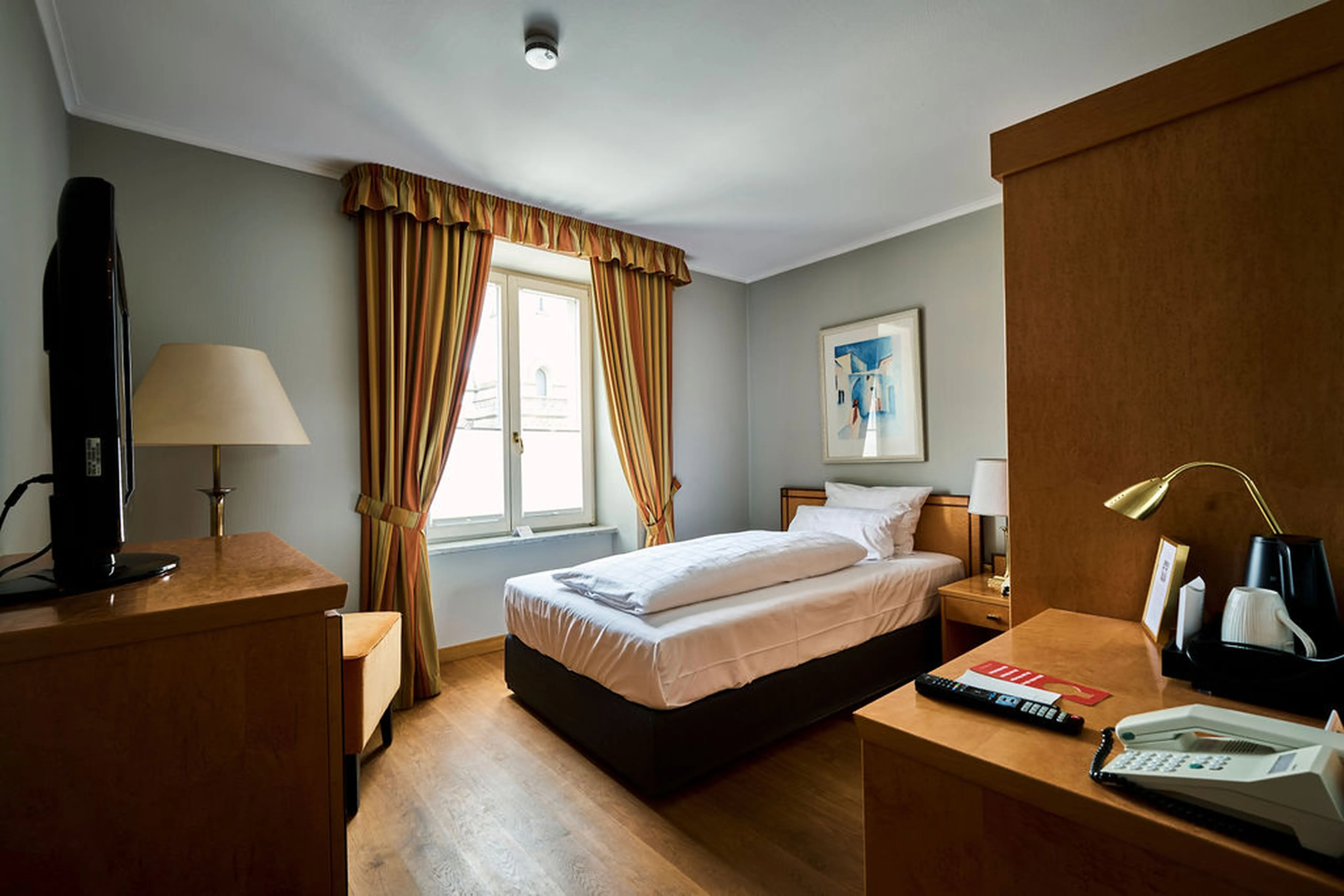 Gemütliches Hotelzimmer mit Einzelbett, Holzmöbeln, Tischlampe, Telefon und TV. Fenster mit gestreiften Vorhängen und einem Bild an der Wand.