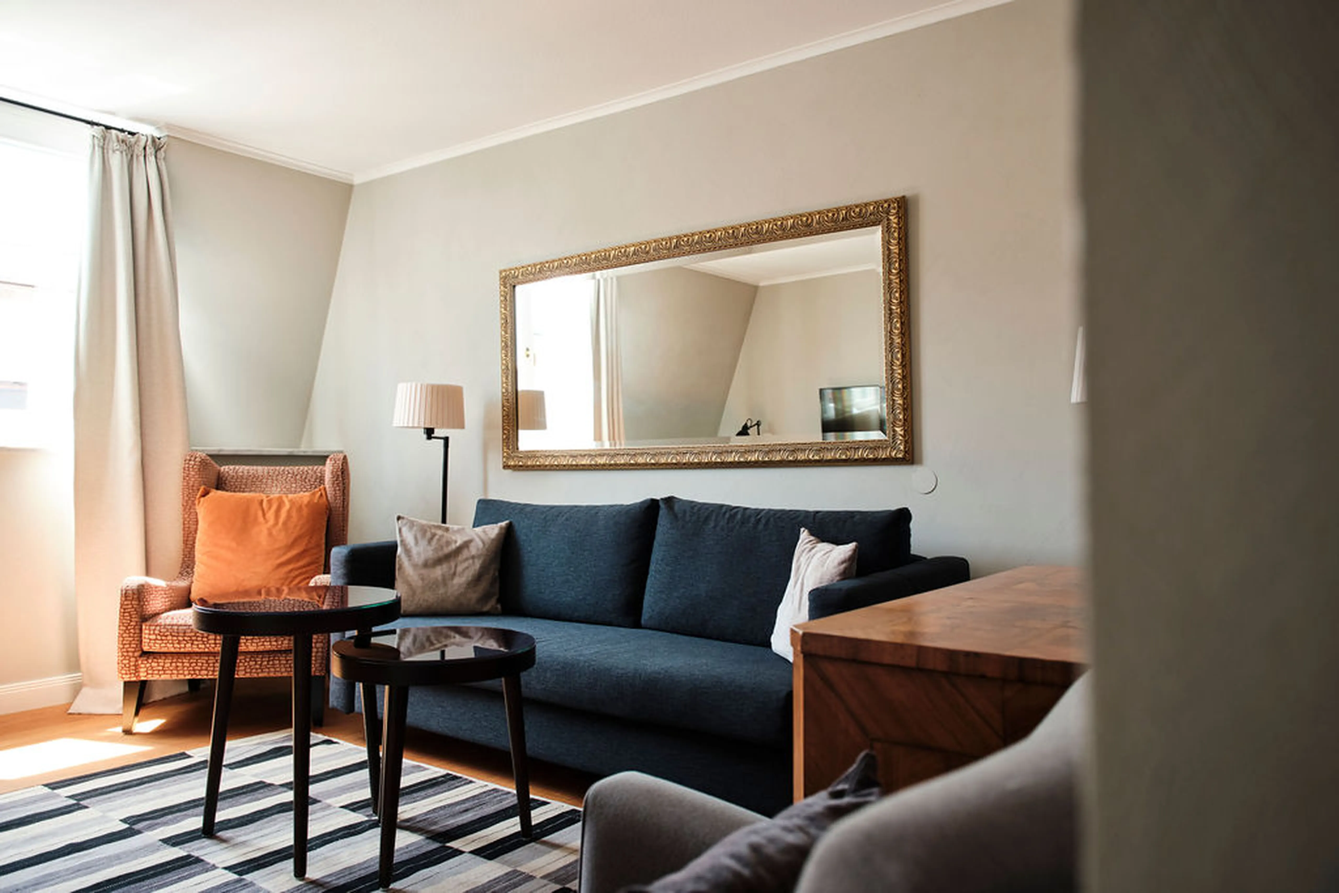 Elegantes Wohnzimmer mit blauer Couch, orangem Sessel, Beistelltischen, großem Spiegel an der Wand und gestreiftem Teppich auf Holzboden.