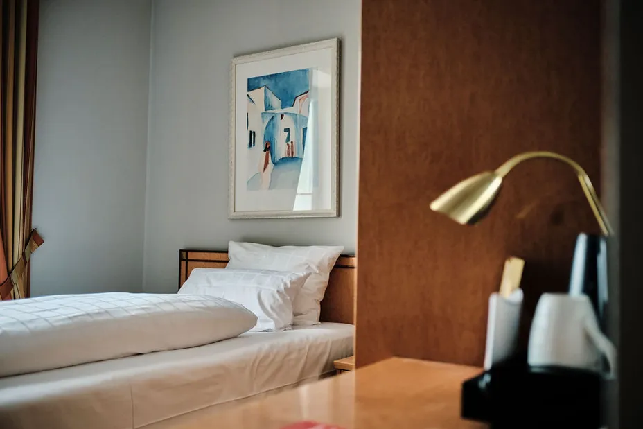 Gemütliches Schlafzimmer mit einem Einzelbett, weißer Bettwäsche, einem gerahmten Bild an der Wand und einer goldenen Tischlampe auf dem Schreibtisch.