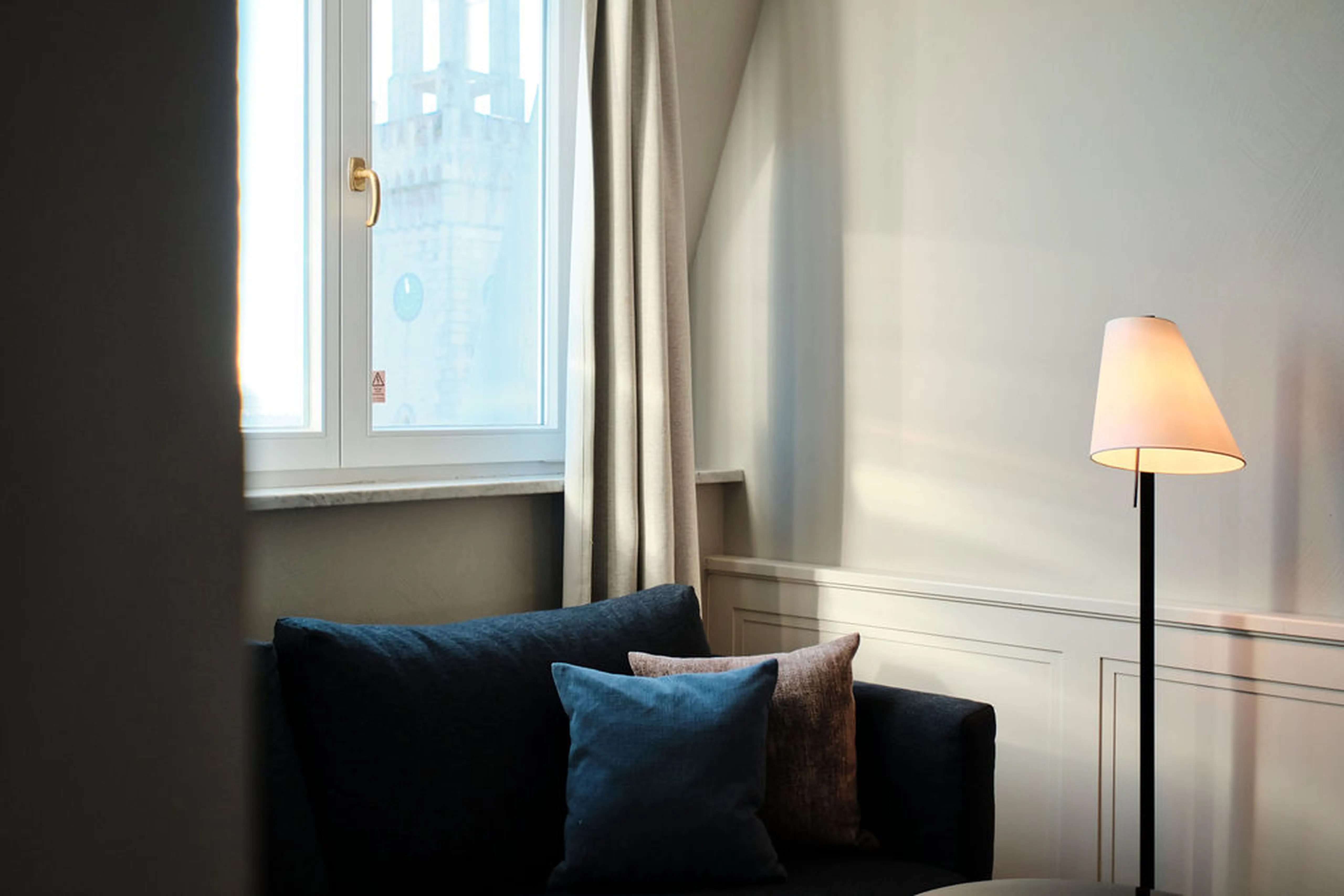 Ein gemütliches Zimmer mit einem dunklen Sofa, blauen Kissen, Stehlampe und einem Fenster mit schlichtem Vorhang, durch das Sonnenlicht fällt.