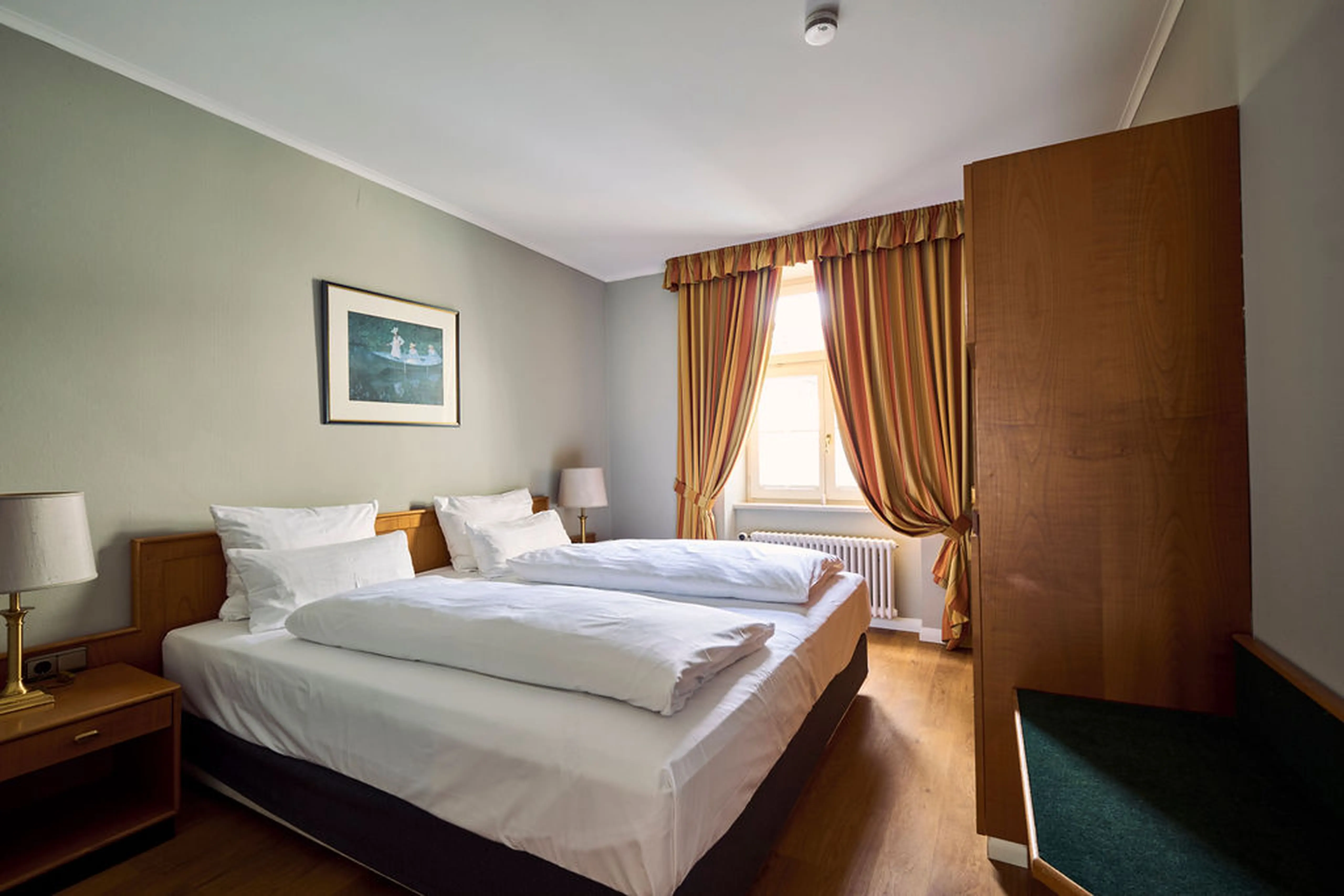Ein schlichtes Hotelzimmer mit Doppelbett, zwei Nachttischen, Vorhängen, Gemälde an der Wand und Tageslicht durch ein Fenster.