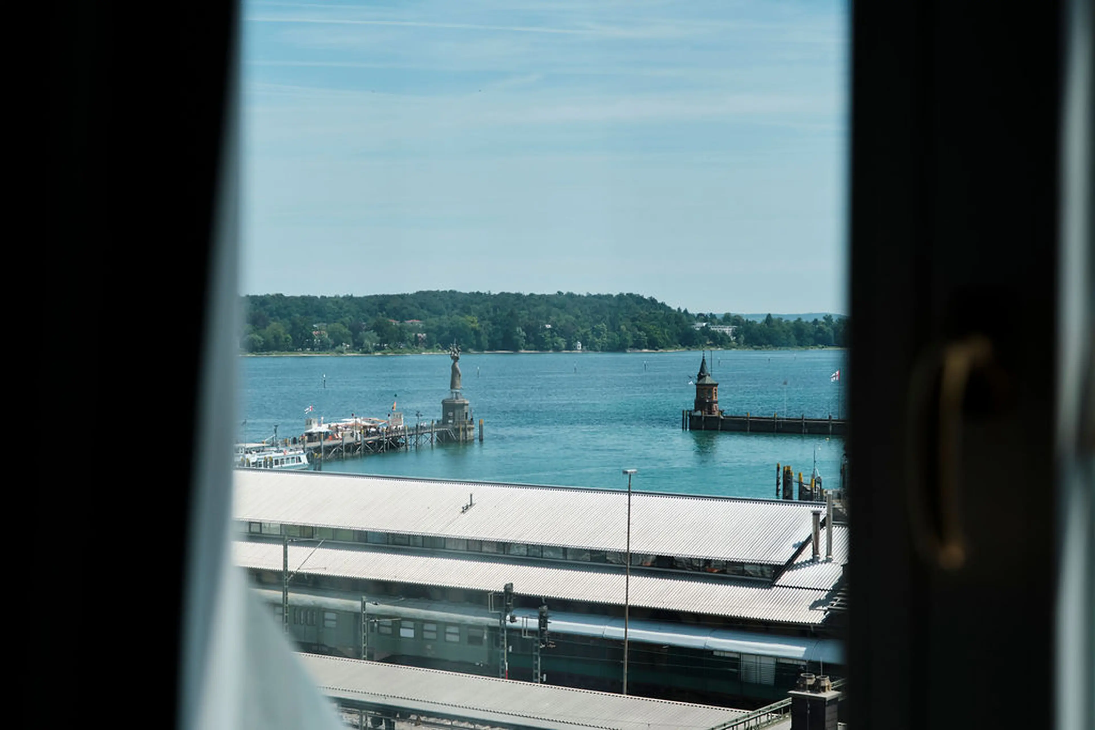 Blick aus einem Fenster auf einen See, die Imperia auf einer Mole und eine umliegende grüne Landschaft bei klarem Himmel.