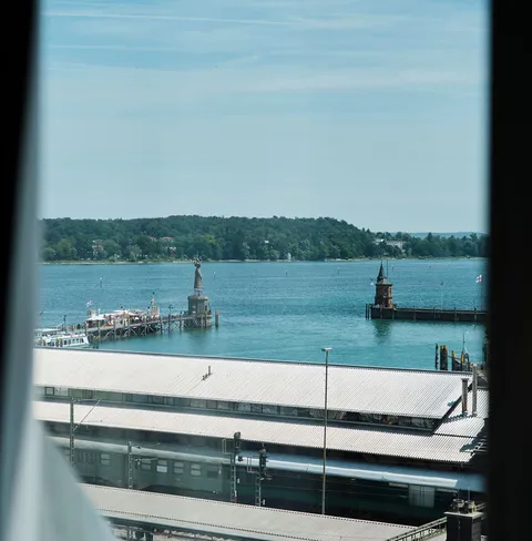 Blick aus einem Fenster auf einen See, die Imperia auf einer Mole und eine umliegende grüne Landschaft bei klarem Himmel.