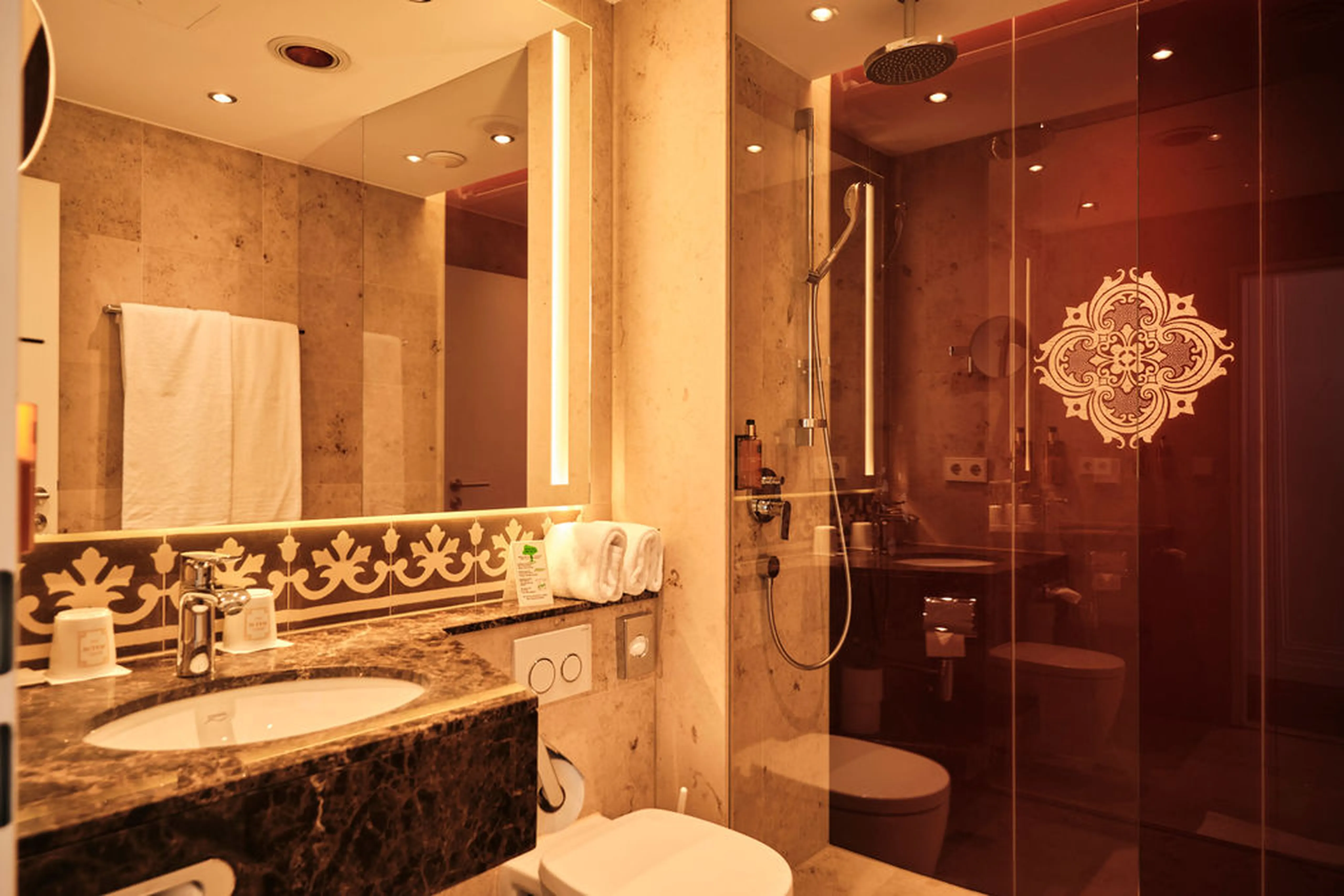 Modernes Badezimmer mit Marmordekor, großer Spiegel, Dusche mit Glaswand und dekorativem Muster, Waschbecken, Handtüchern und Beleuchtung.