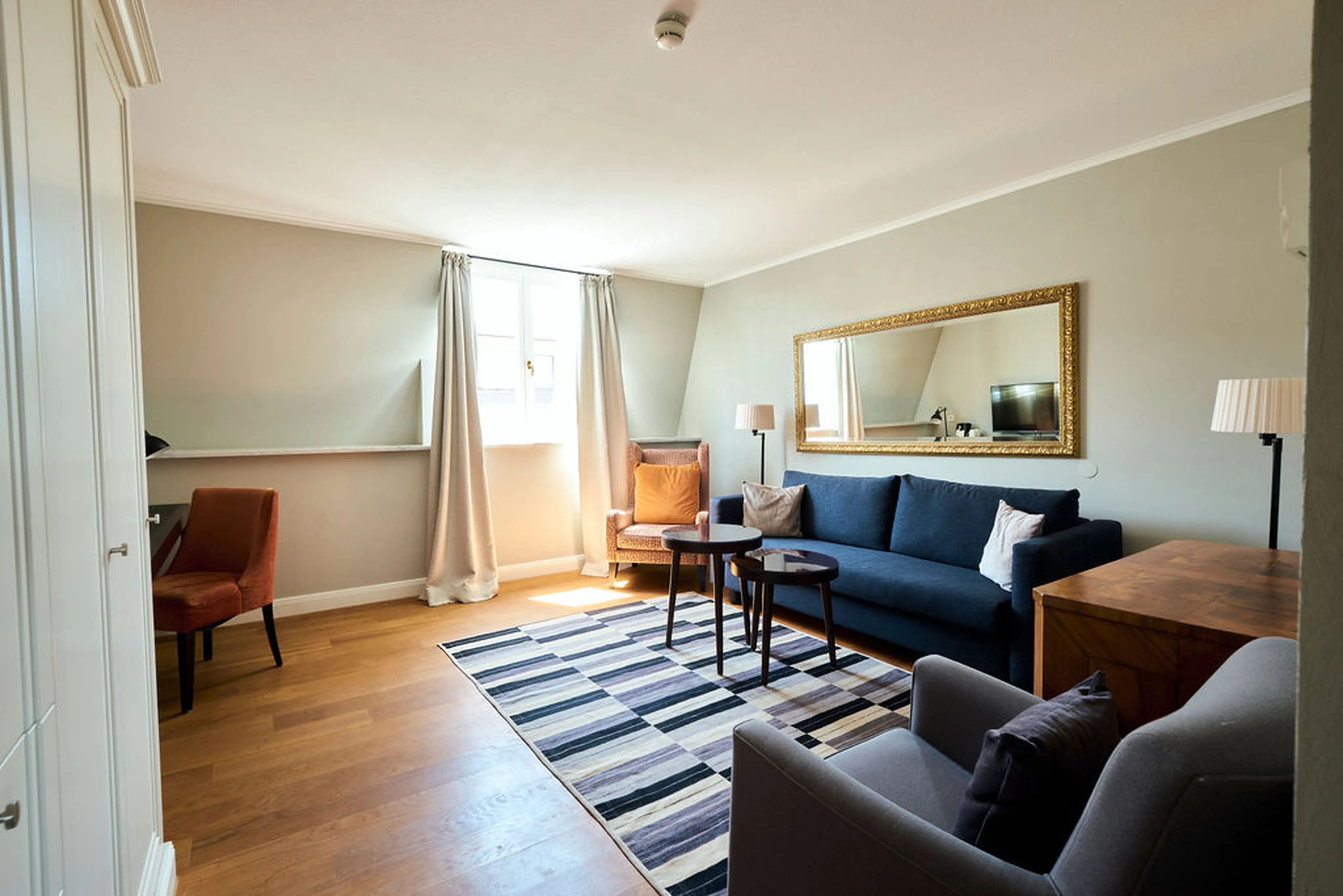 Wohnzimmer mit blauer Couch, grauem Sessel, gestreiftem Teppich, großem Spiegel und zwei Fenstern, die Tageslicht einlassen.