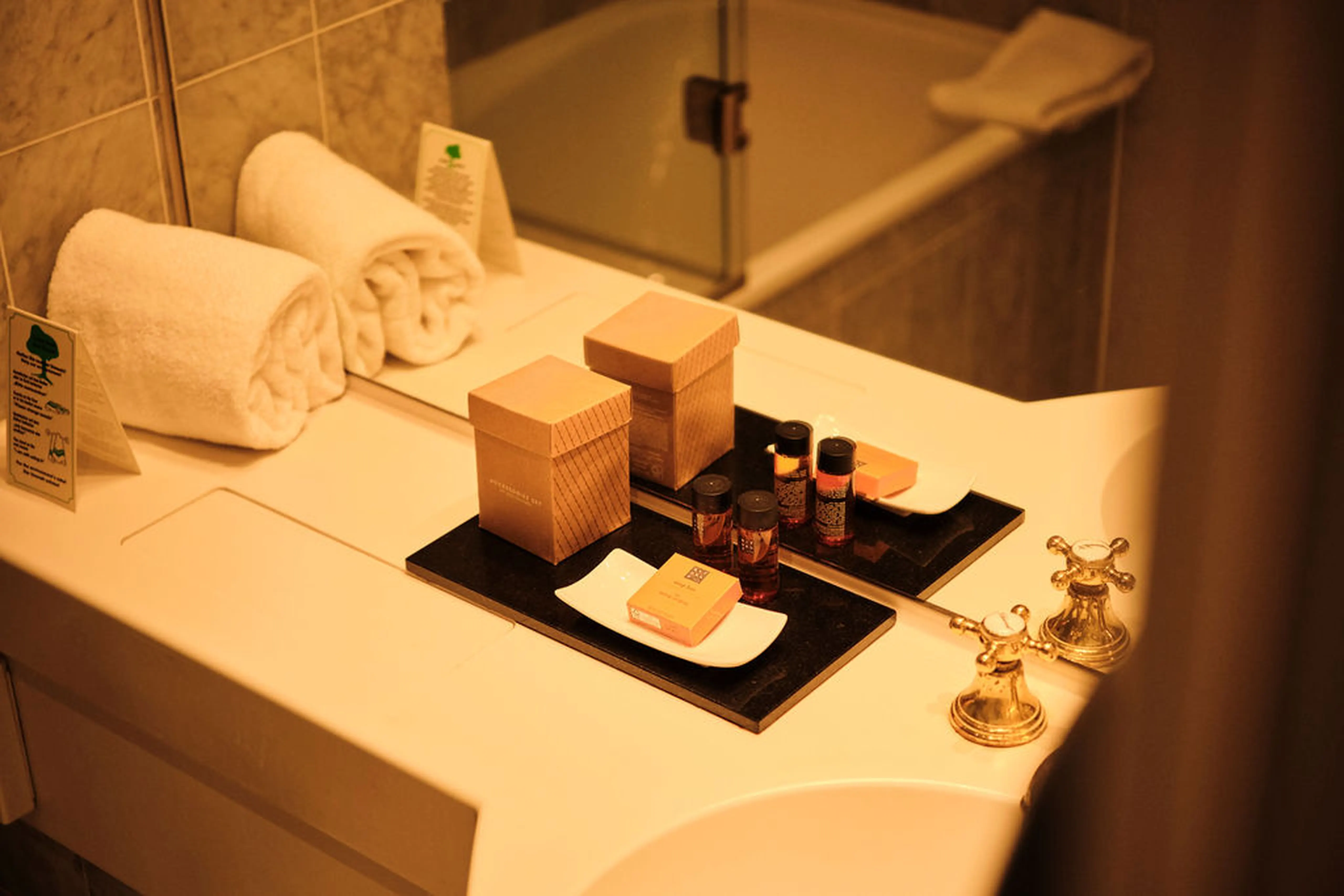 Auf einem Badezimmerwaschtisch stehen zwei Handtücher, Pflegeprodukte, Seife und eine braune Schachtel neben goldenen Wasserhähnen.