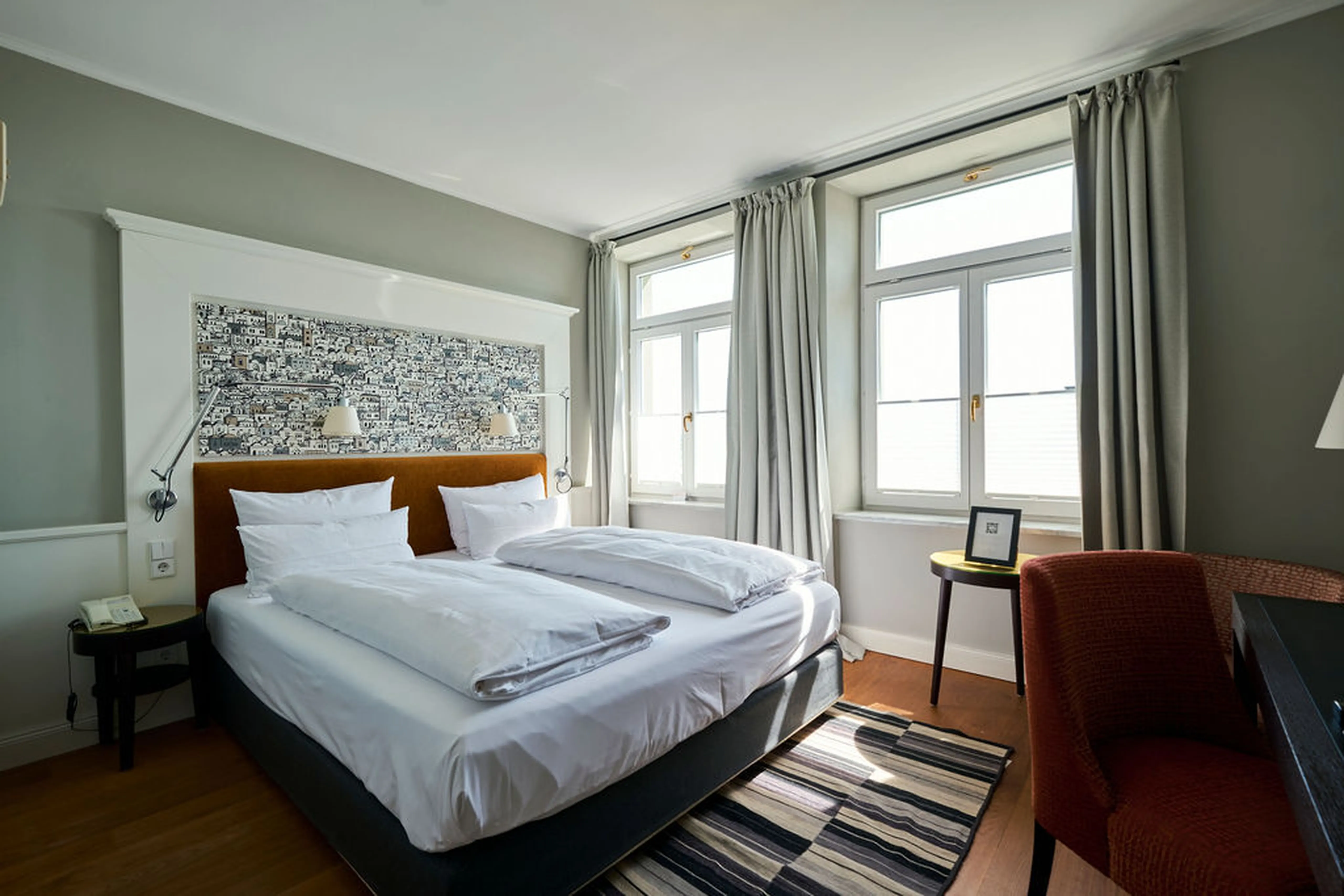 Helles Hotelzimmer mit Doppelbett, modernem Kunstwerk am Kopfteil, zwei Fenstern mit grauen Vorhängen, rotem Sessel und Schreibtisch auf Holzboden.