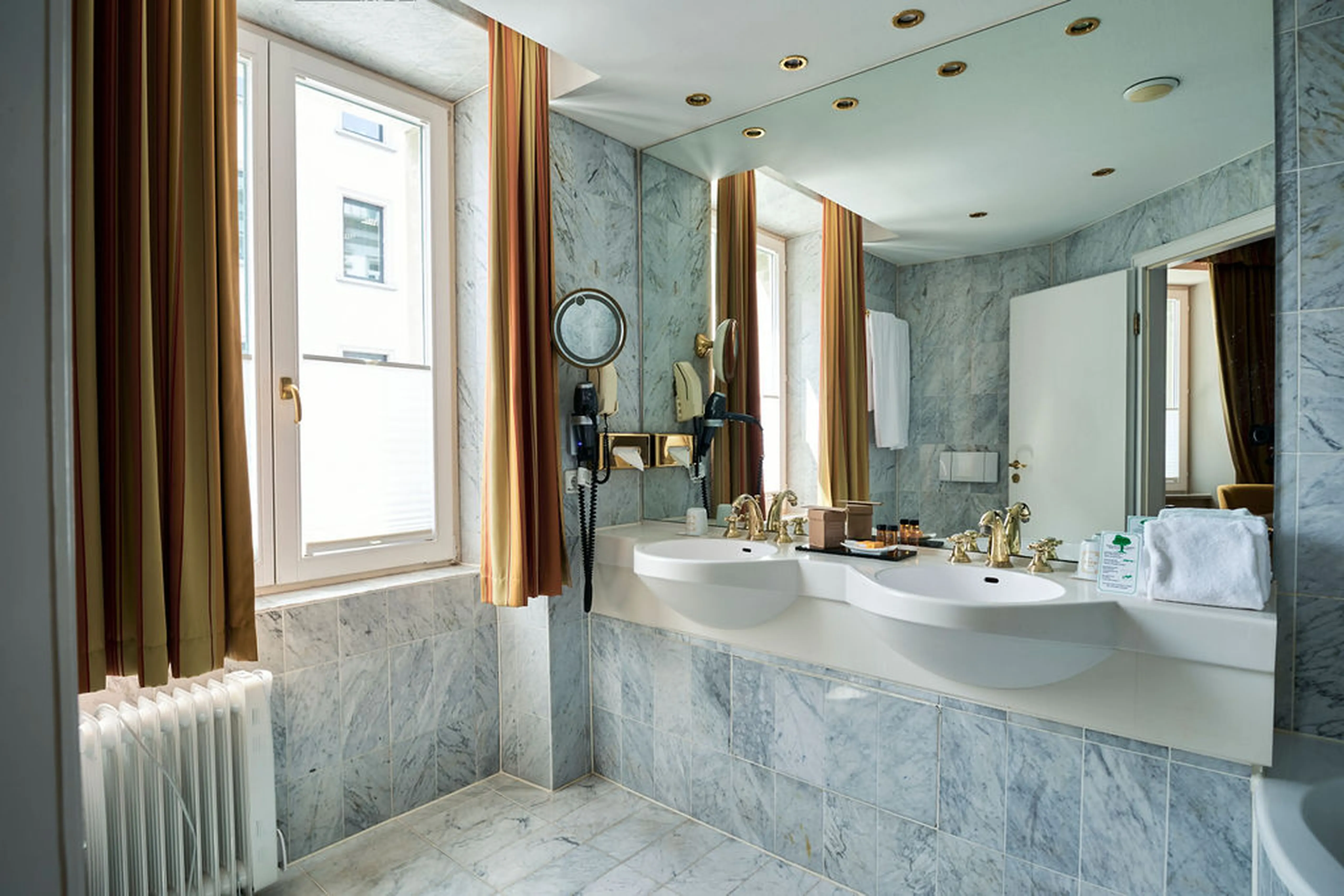 Modernes Badezimmer mit Marmorverkleidung, zwei Waschbecken, goldenen Wasserhähnen, großen Spiegeln, Fenstern mit Vorhängen und Handtüchern.