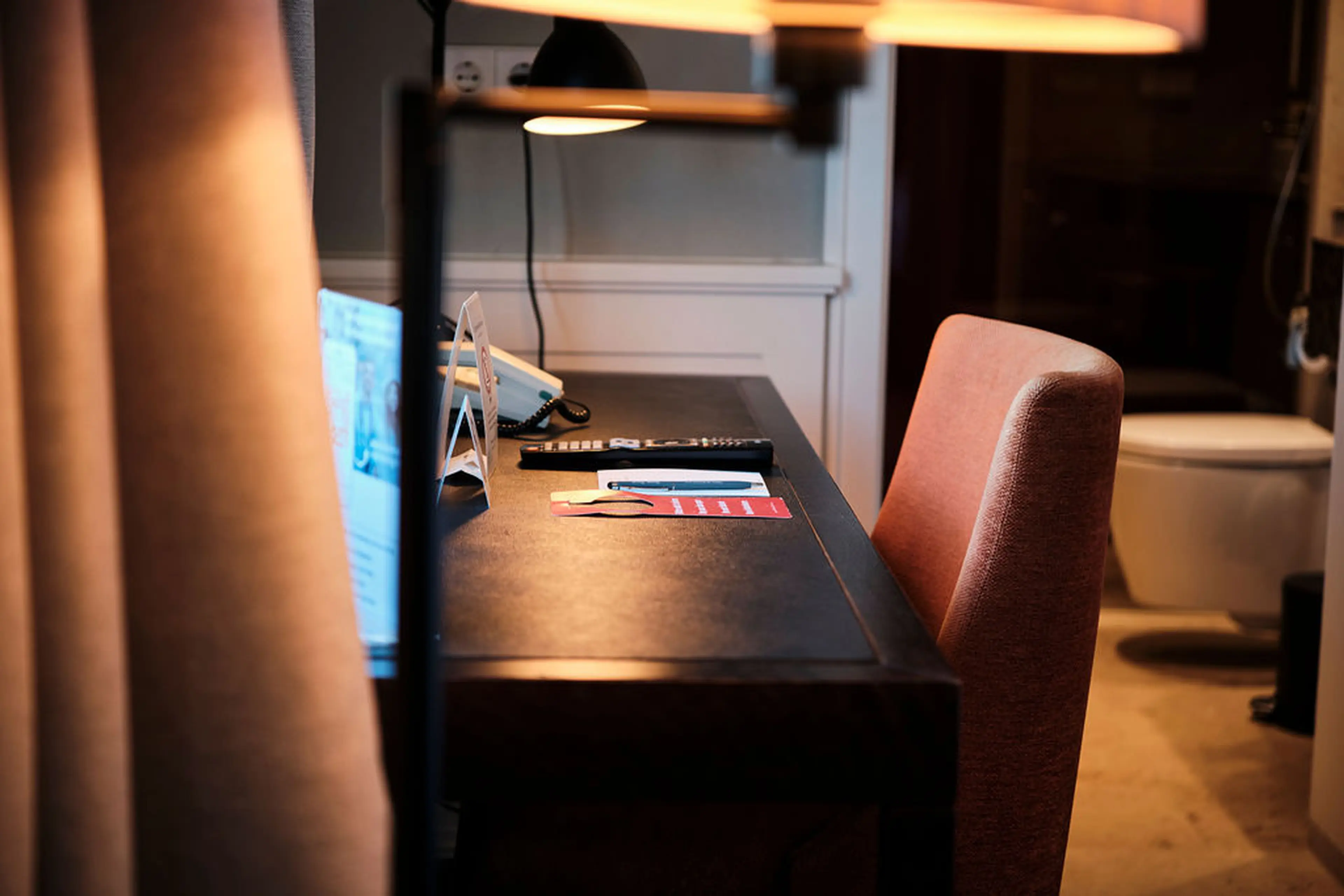 Ein Schreibtisch mit Lampe und Stuhl in einem Hotelzimmer, darauf liegen ein Telefon, eine Fernbedienung und Informationsmaterial.
