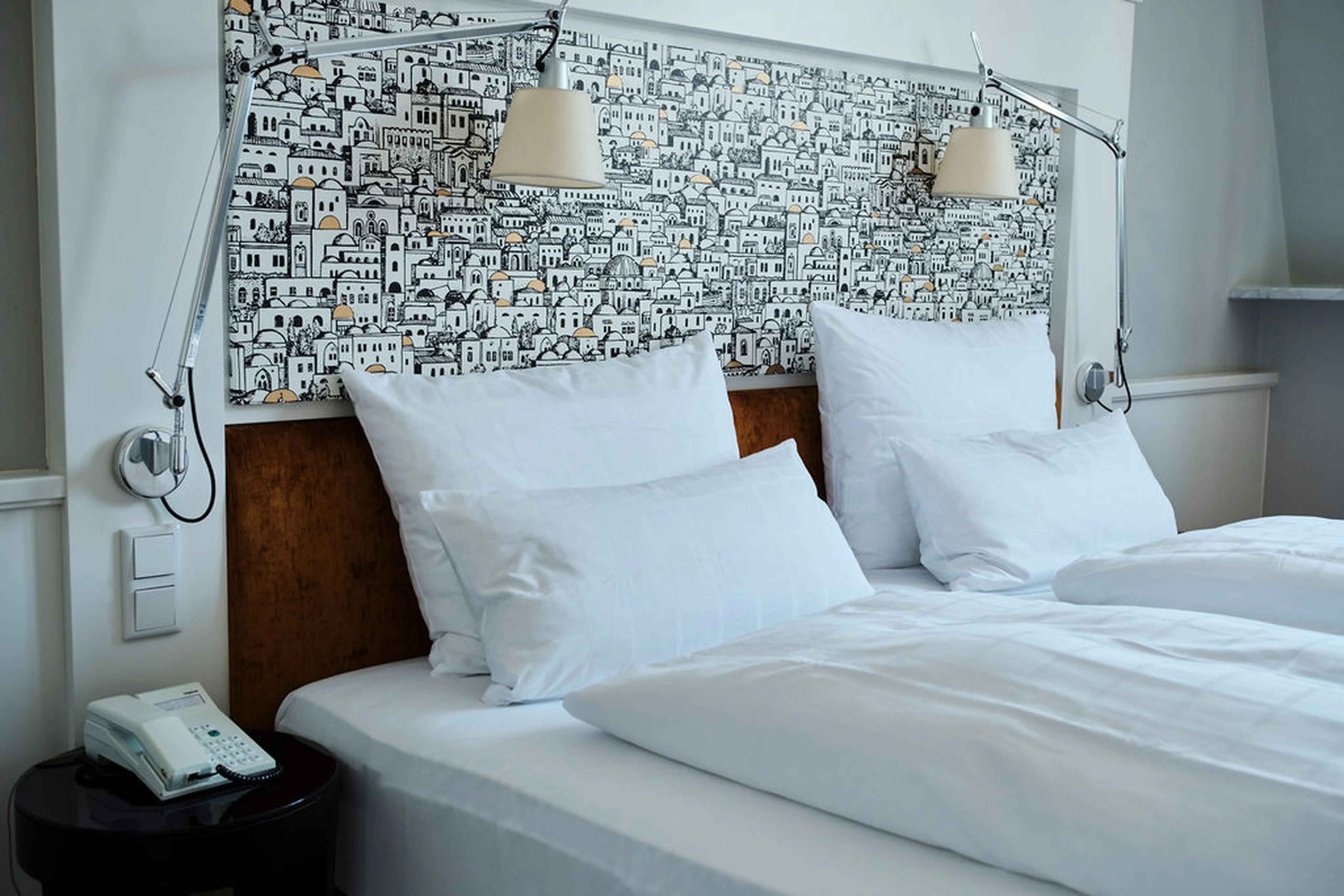 Ein Hotelzimmer mit einem gemütlichen Doppelbett, weißen Kissen, einer dekorativen Kopfleiste mit Stadtmuster und zwei Wandlampen.