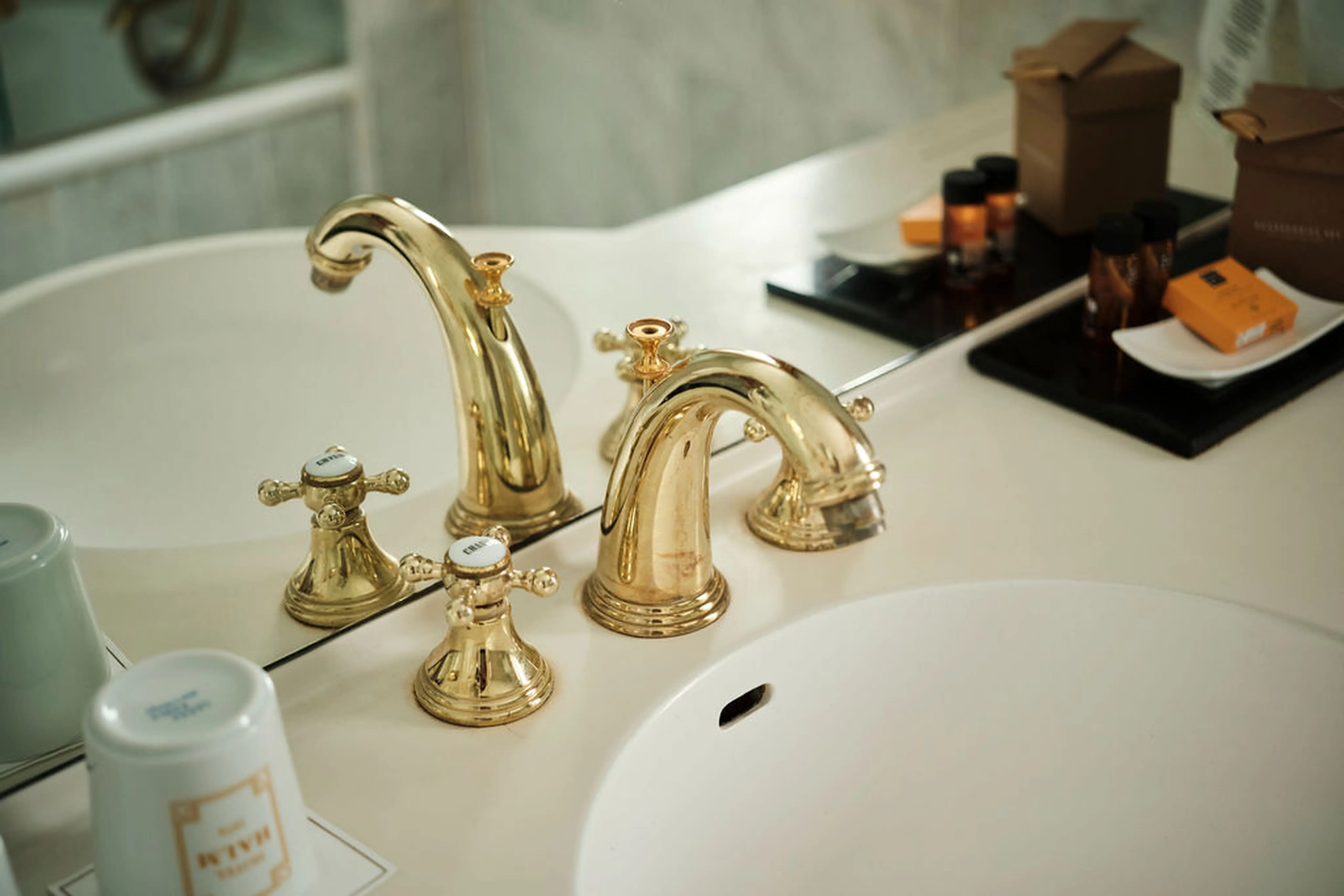 Goldene Wasserhähne auf einem Doppelwaschbecken, umgeben von Toilettenartikeln und kleinen braunen Boxen auf einem weißen Waschtisch.
