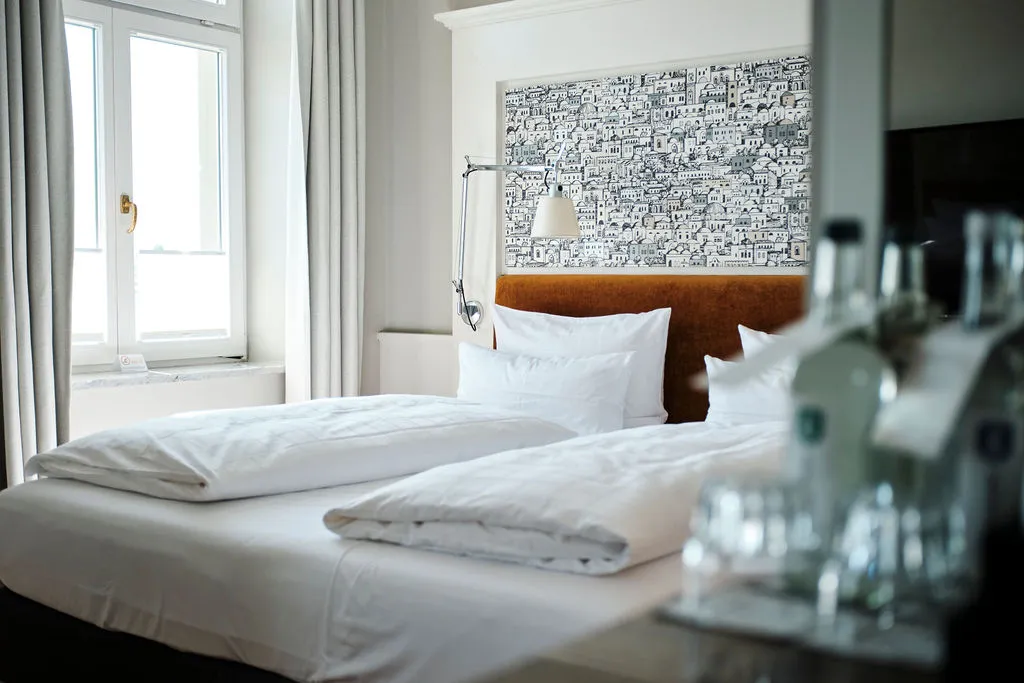 Ein stilvolles Hotelzimmer mit einem großen Bett, weißen Bettbezügen und einem Fenster mit Vorhängen. Im Vordergrund Wasserflaschen.