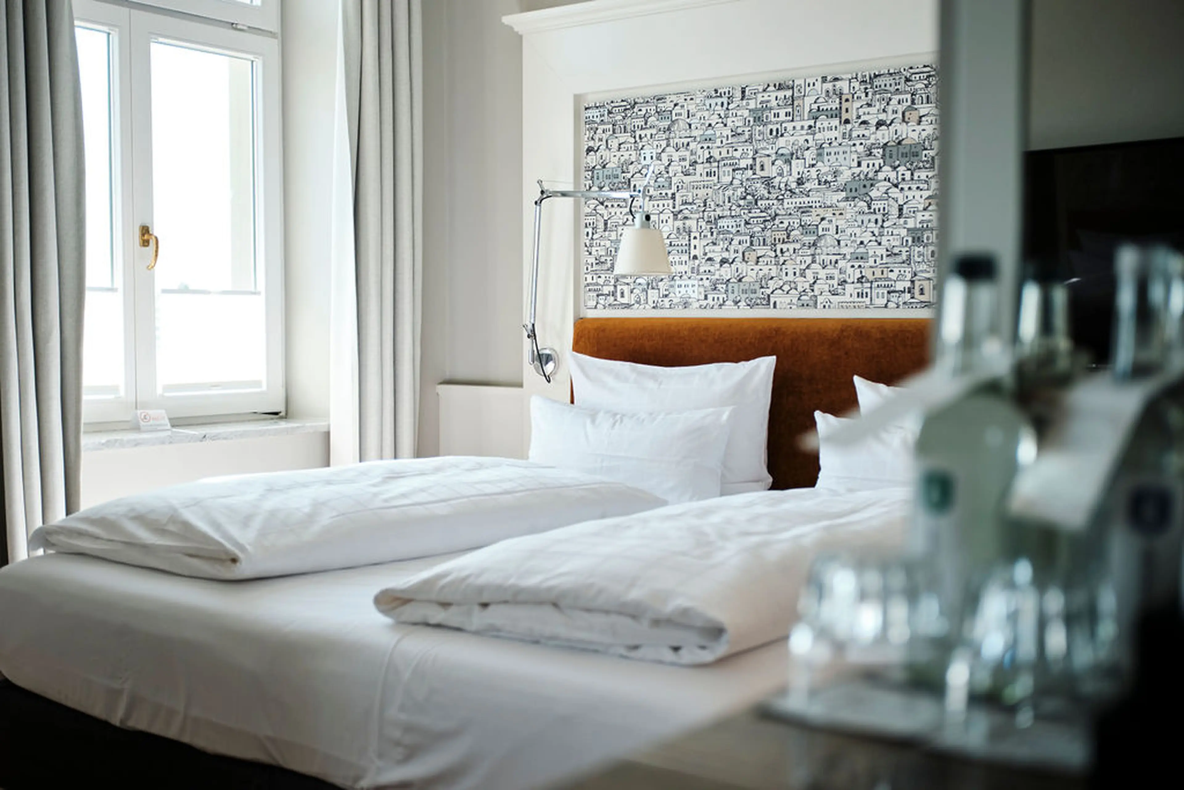 Ein stilvolles Hotelzimmer mit einem großen Bett, weißen Bettbezügen und einem Fenster mit Vorhängen. Im Vordergrund Wasserflaschen.