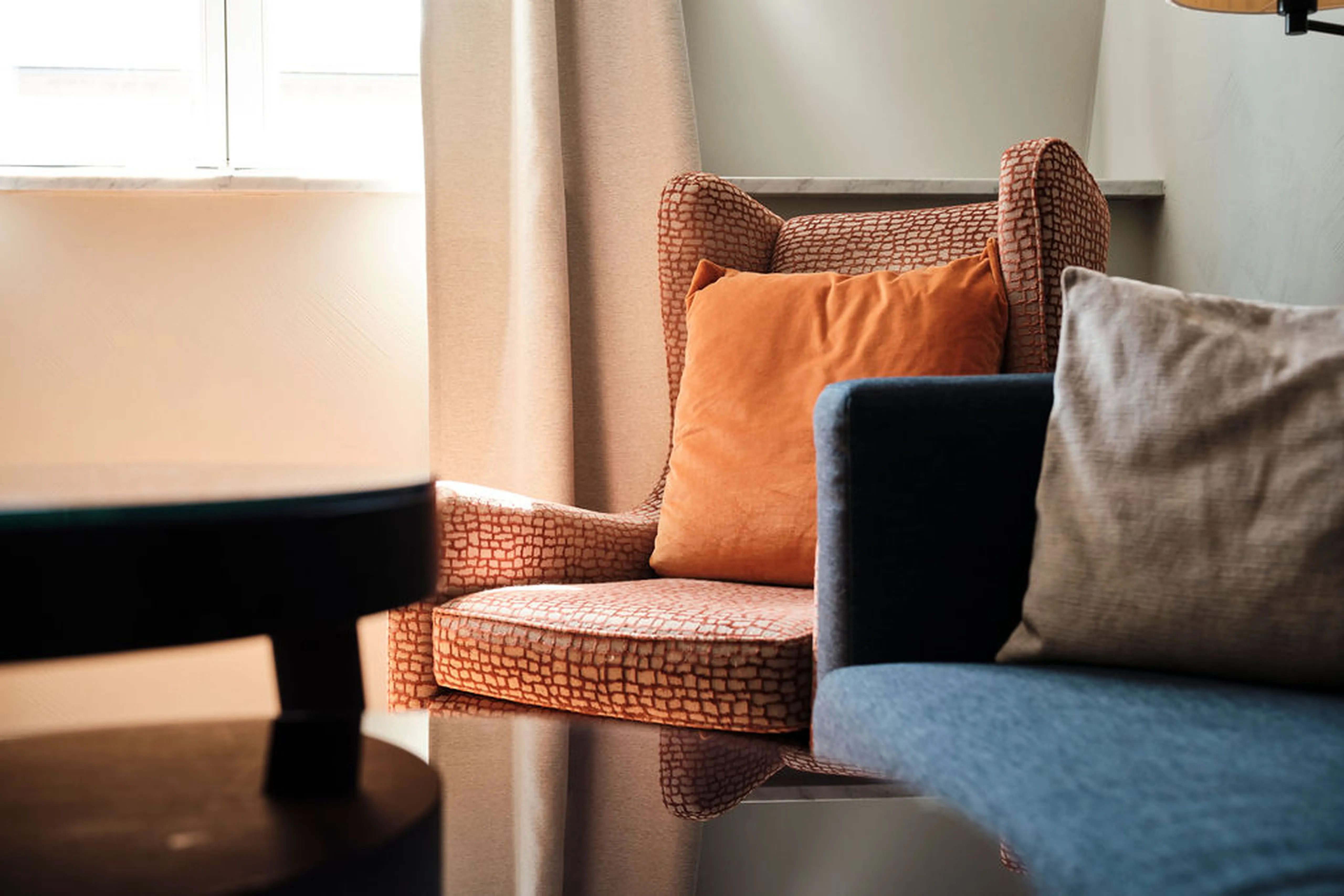 Gemütlicher Sessel mit orangefarbenem Kissen neben einem blauen Sofa, vor einem Fenster mit beigefarbenem Vorhang, beleuchtet von Sonnenlicht.