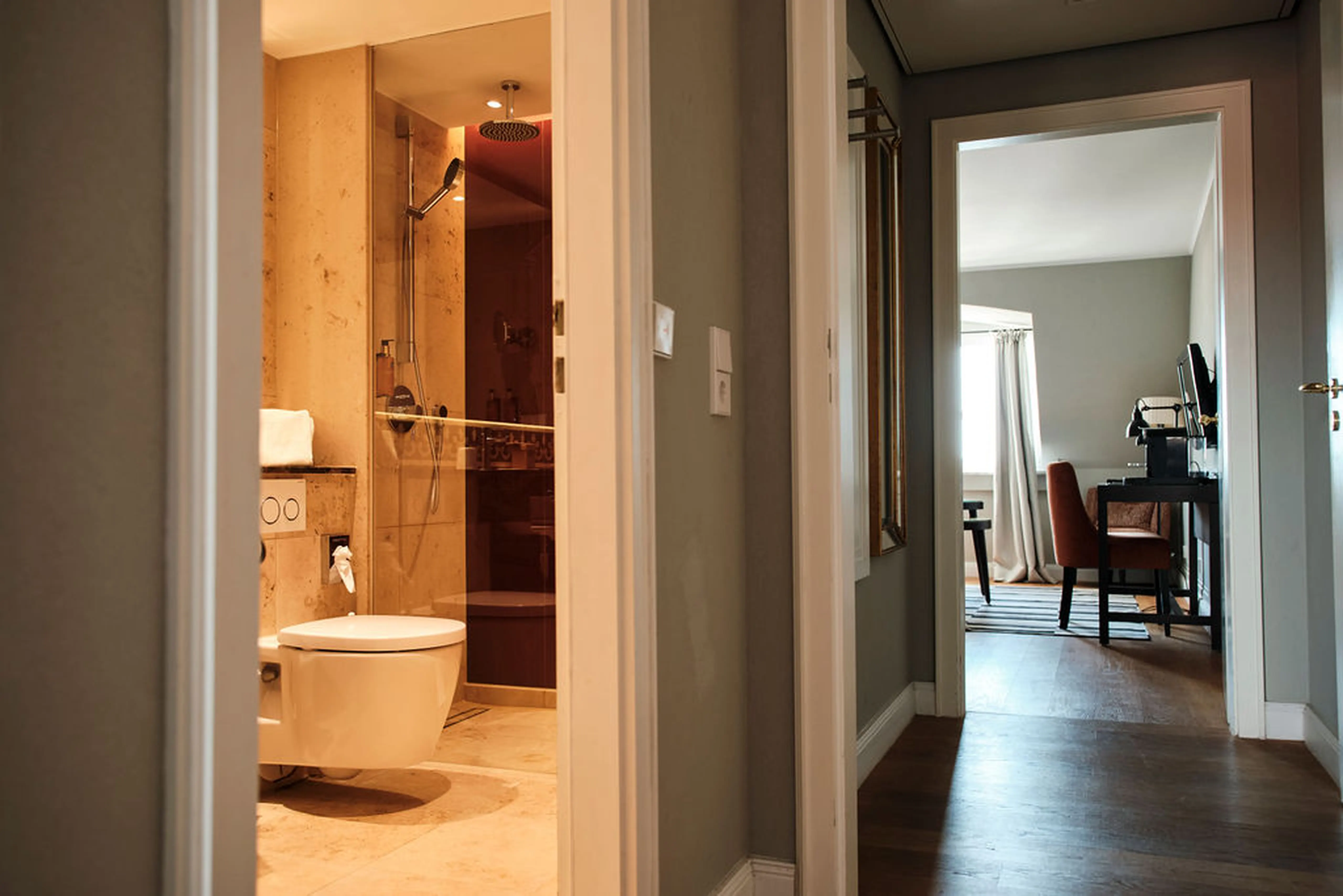 Blick in ein Badezimmer mit moderner Dusche und Toilette, daneben ein Flur, der zu einem Raum mit Schreibtisch und Stuhl führt.