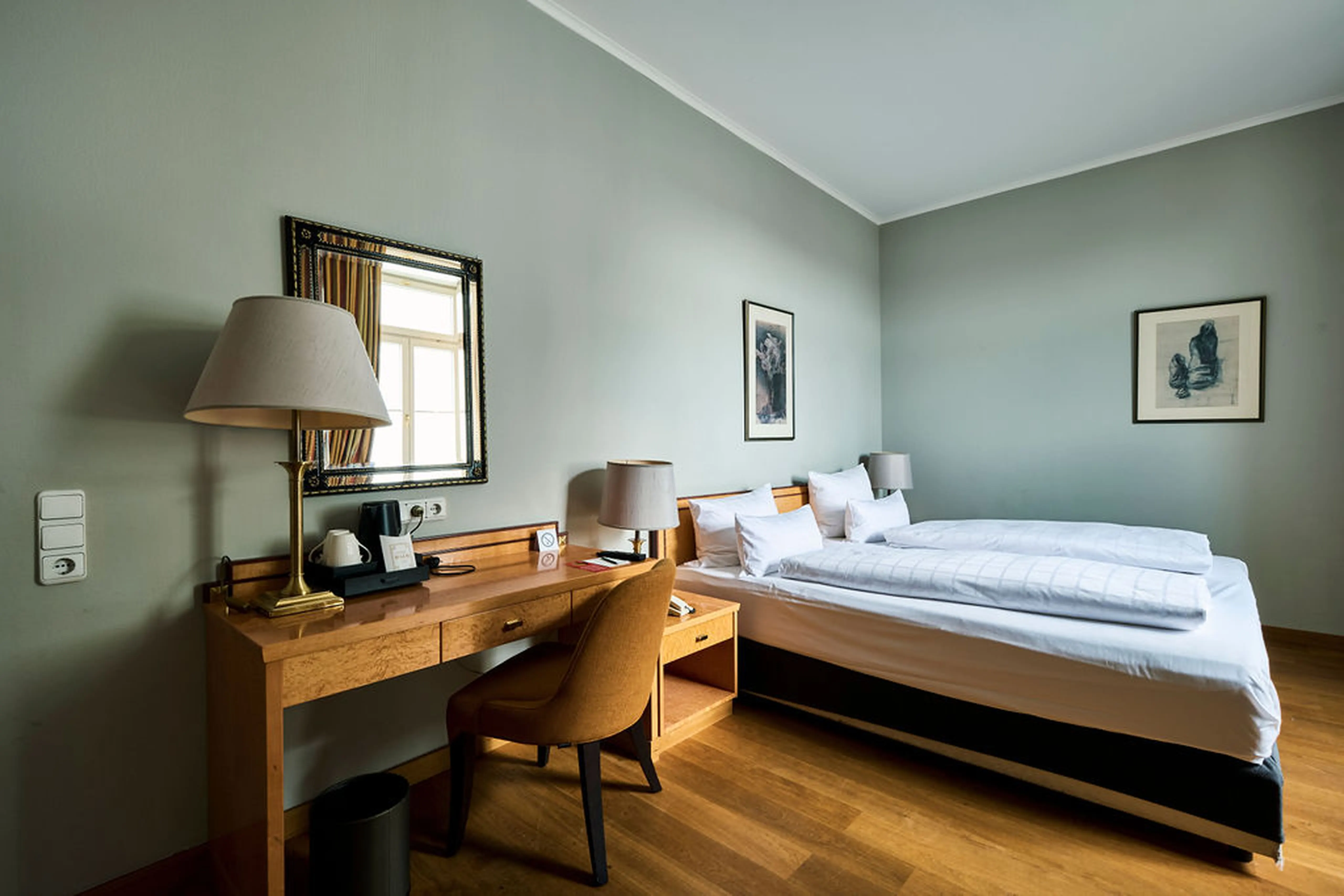 Helles Hotelzimmer mit Doppelbett, Schreibtisch mit Lampe und Kaffeemaschine. Bilder an der Wand und ein Fenster mit Tageslicht.