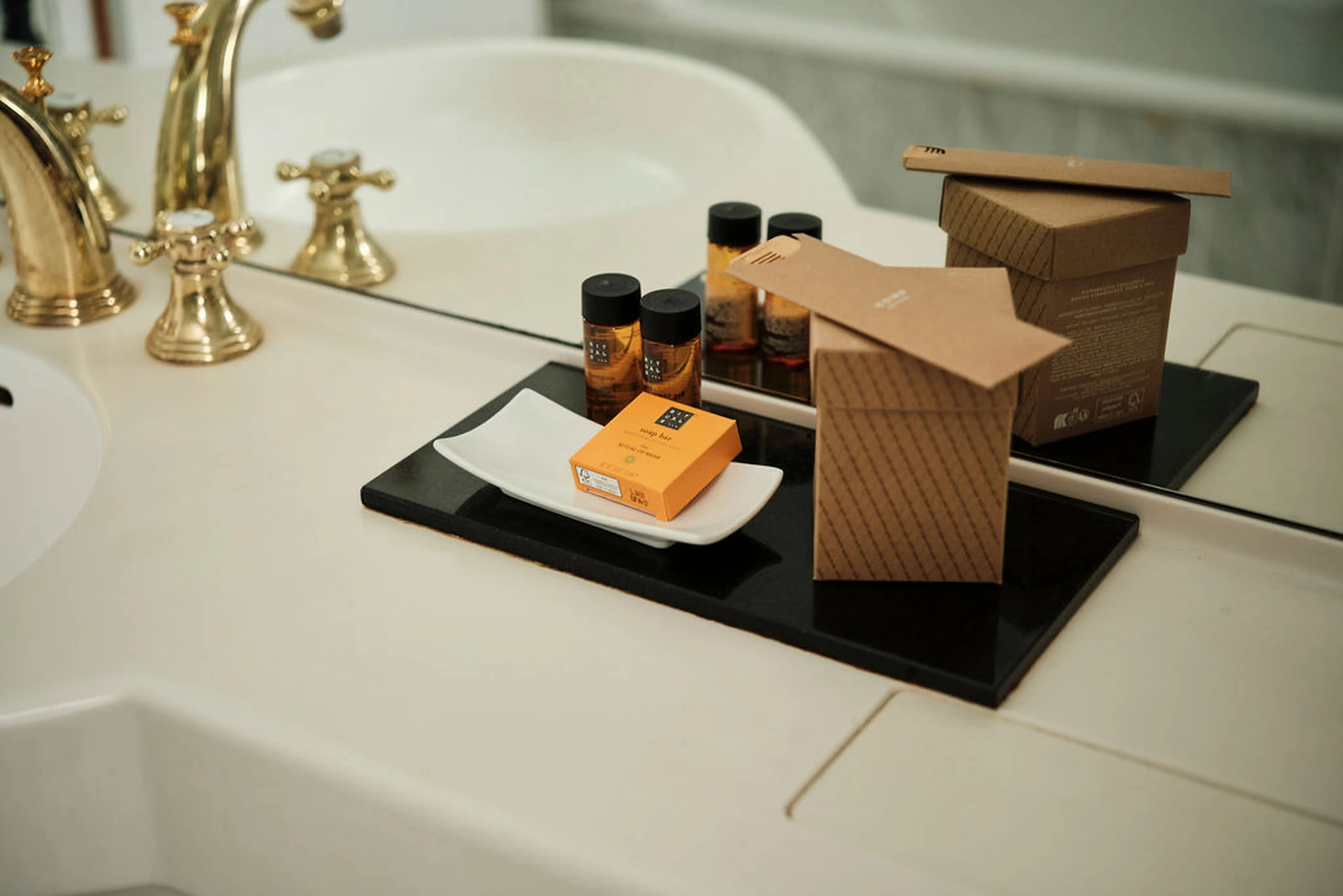 Premium-Badeartikel und Verpackungen auf einem Tablett, neben einem weißen Waschbecken mit goldenen Wasserhähnen.