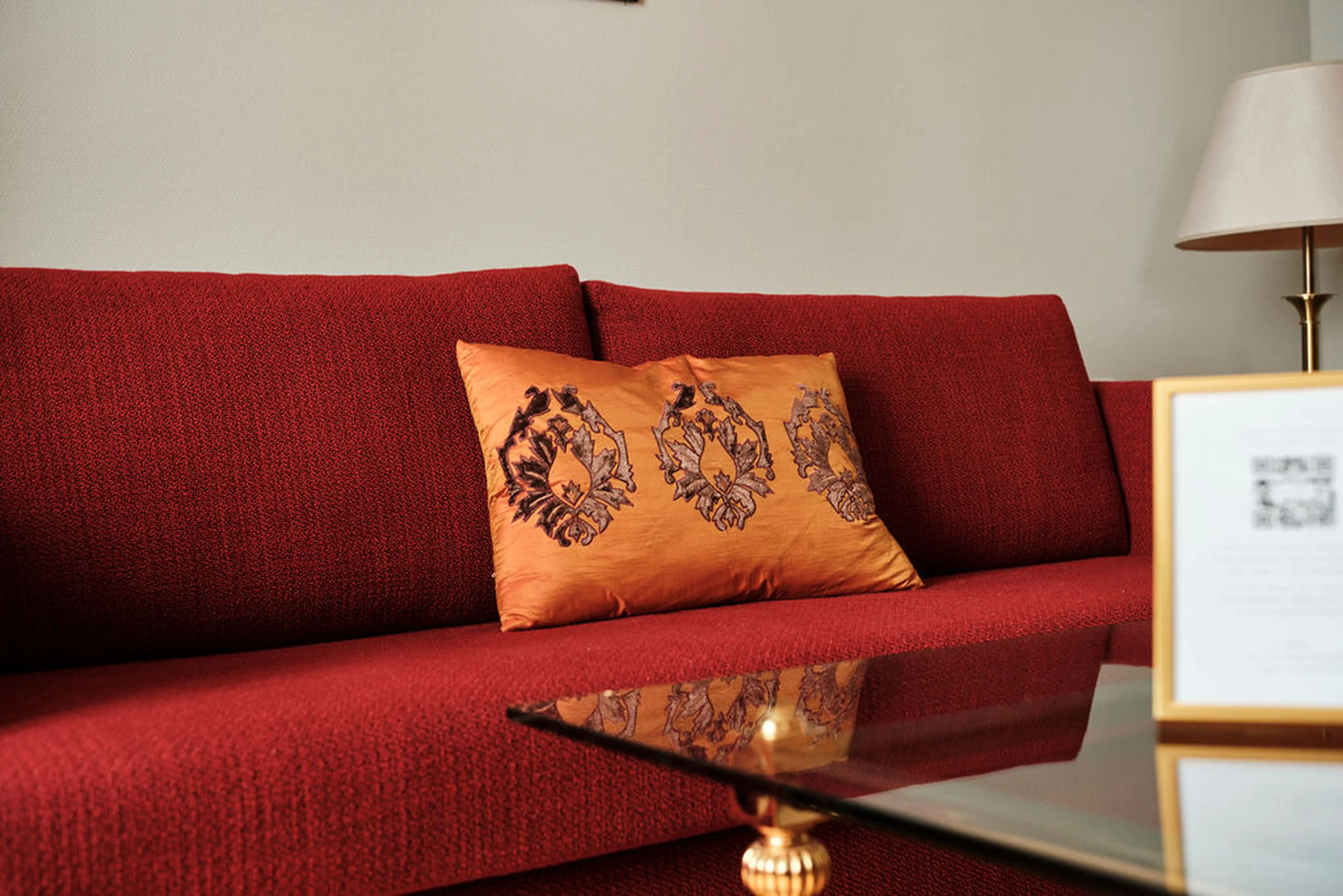 Rote Couch mit einem orange verzierten Kissen, daneben ein Glastisch mit goldenen Details und eine Tischlampe im Hintergrund.