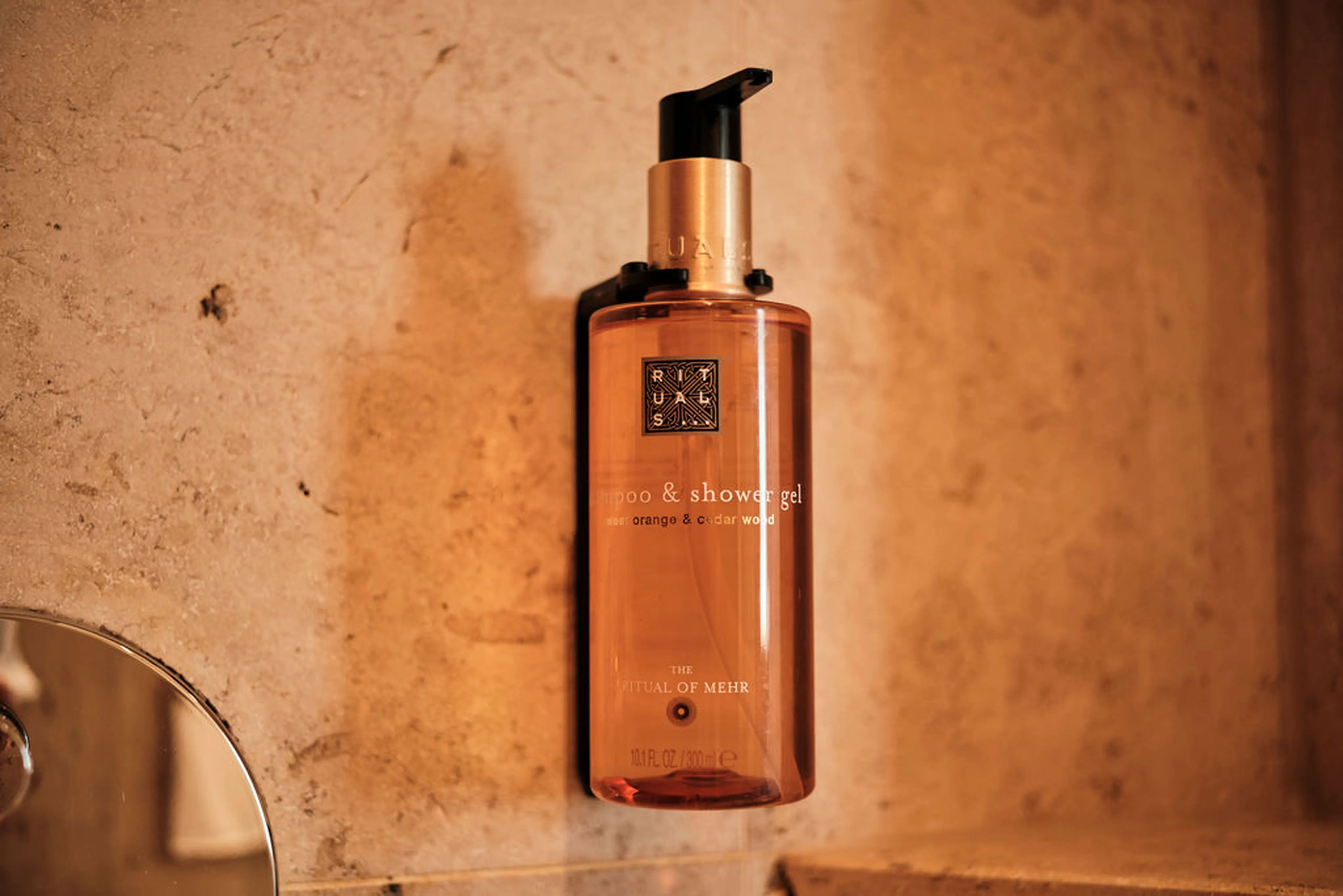 Amberfarbene Flasche mit Rituals Shampoo und Duschgel, montiert an einer beigen Wand, mit Spender an der Oberseite.