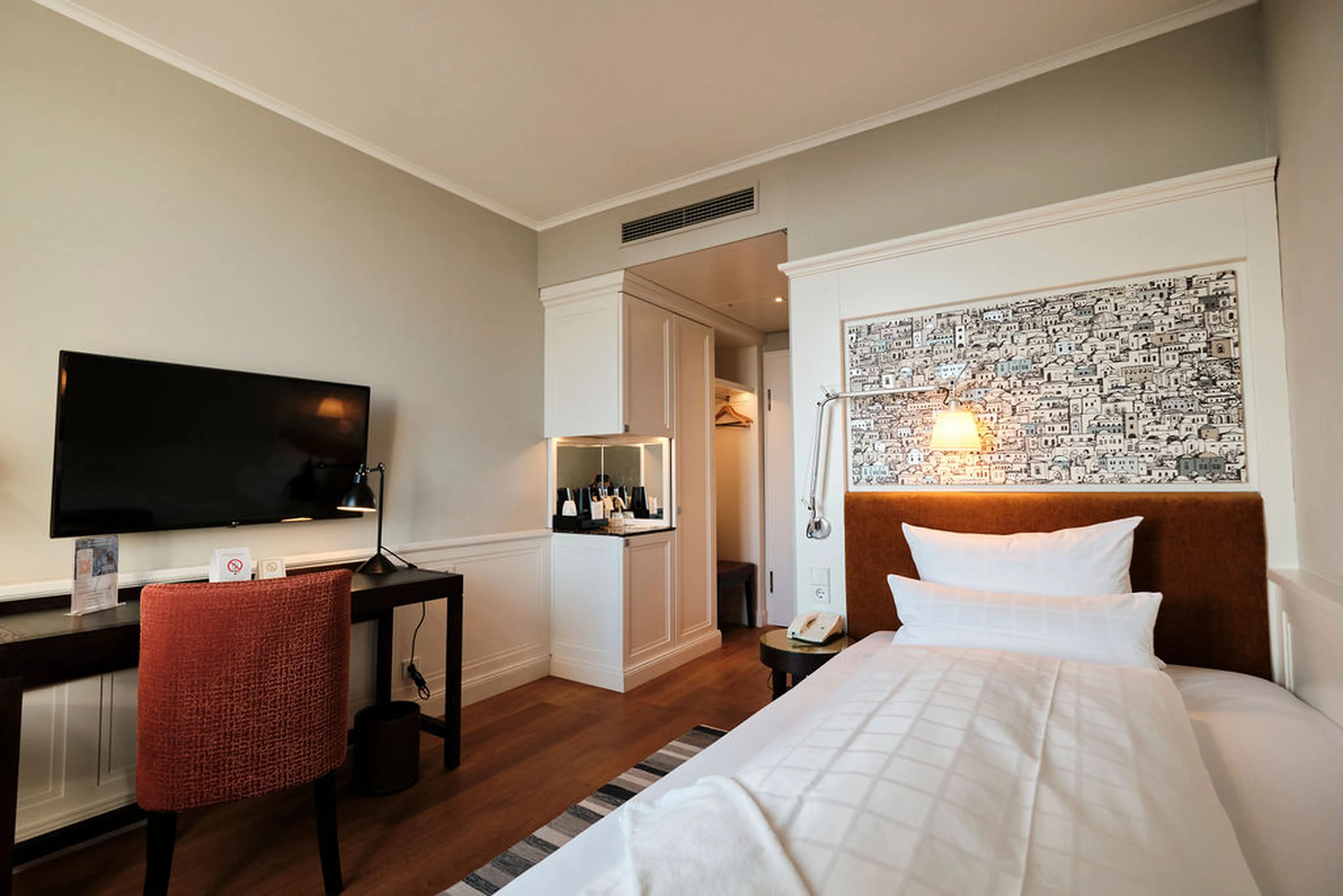 Modernes Hotelzimmer mit Einzelbett, Wandlampe, TV, Schreibtisch, rotem Stuhl, Telefon und kleinem Kleiderschrank. Wanddekor über dem Bett.