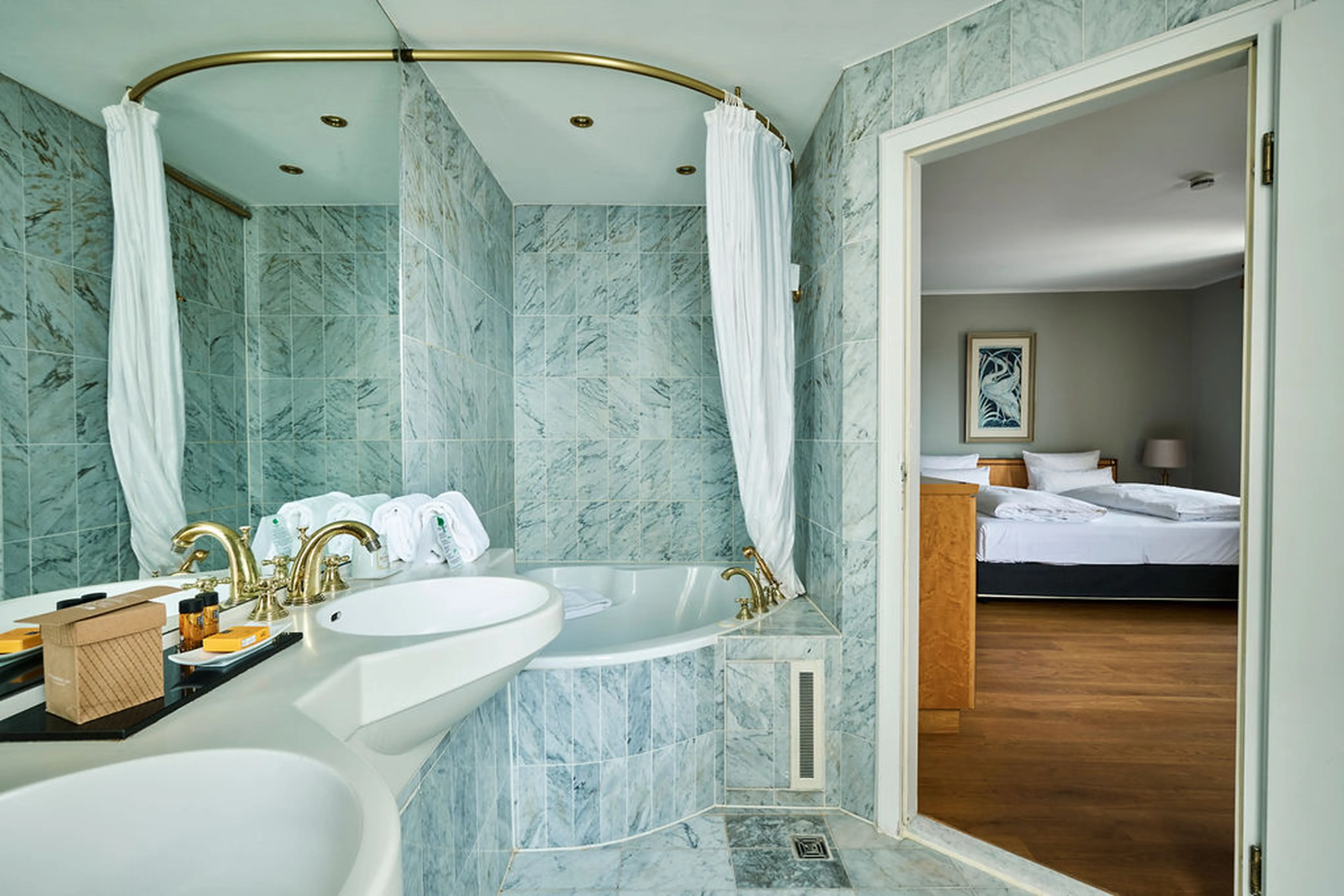 Luxuriöses Badezimmer mit Marmorfliessen, Doppelwaschbecken, goldenen Armaturen und Badewanne. Geöffnetes Fenster zum Schlafzimmer mit großem Bett.