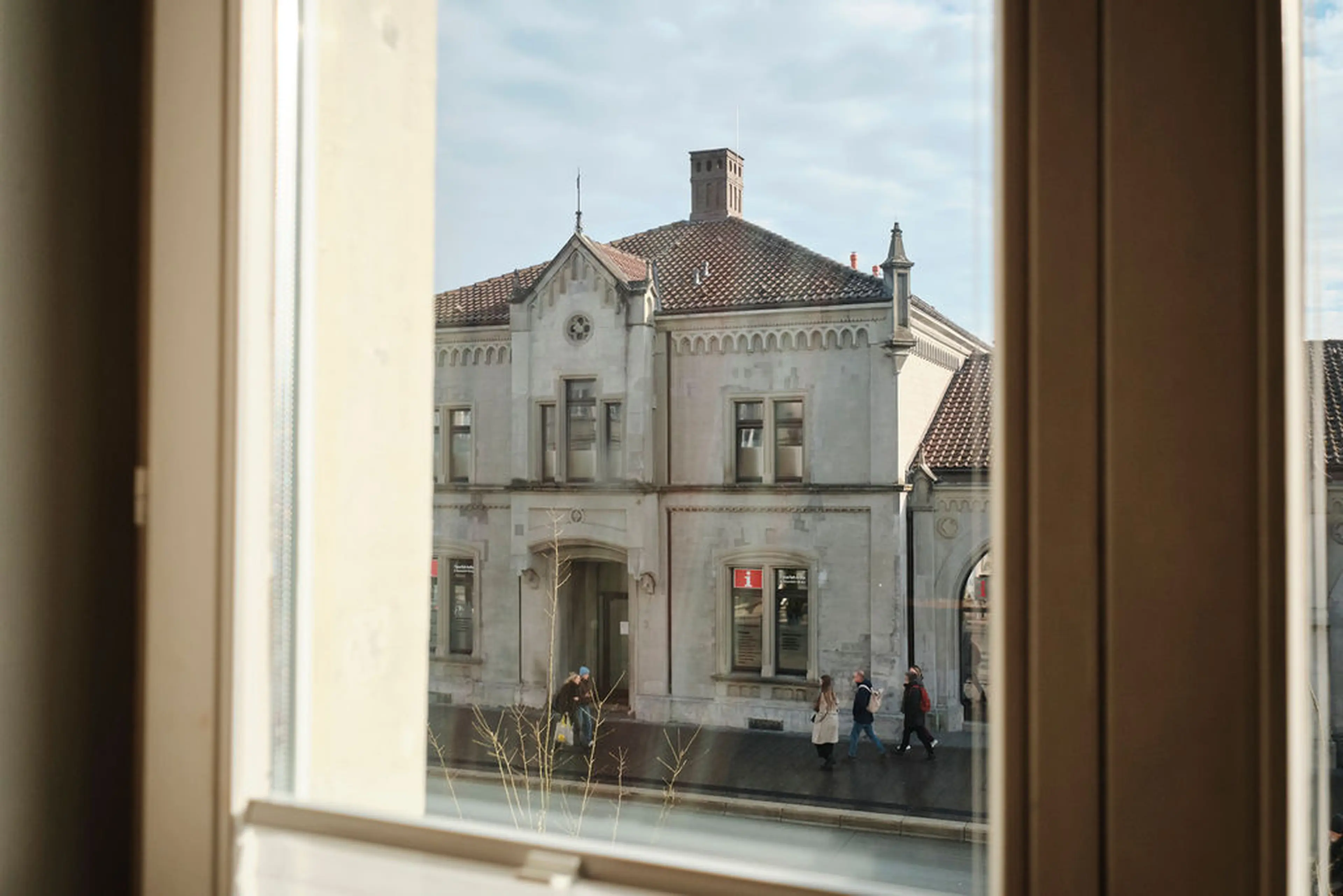Blick aus einem Fenster auf ein historisches Gebäude mit roten Ziegeldächern, Menschen gehen auf dem Bürgersteig entlang.