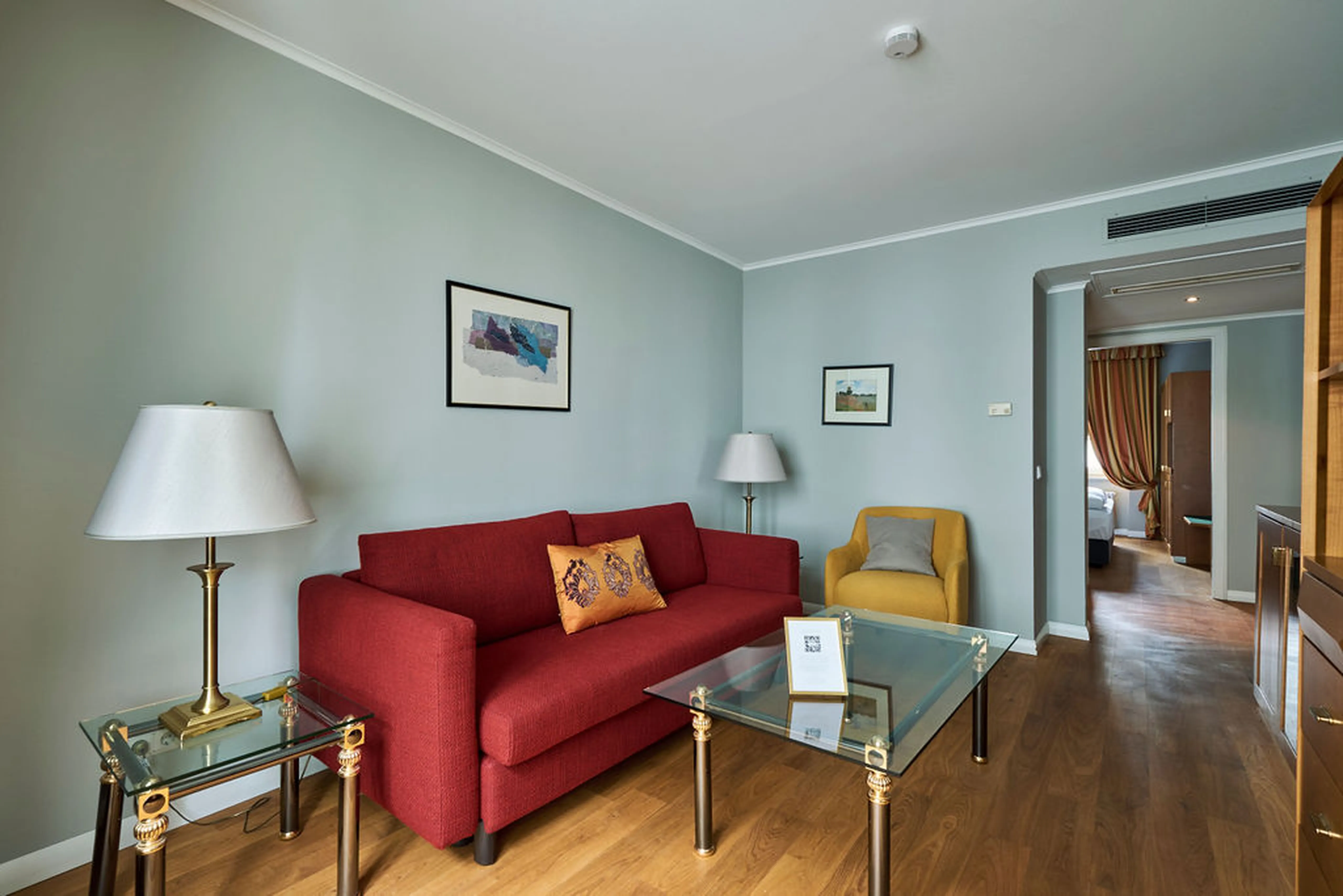 Wohnzimmer mit rotem Sofa, gelbem Sessel, zwei Tischlampen, Glastisch und Wandbildern. Holzboden führt zu einem angrenzenden Raum.