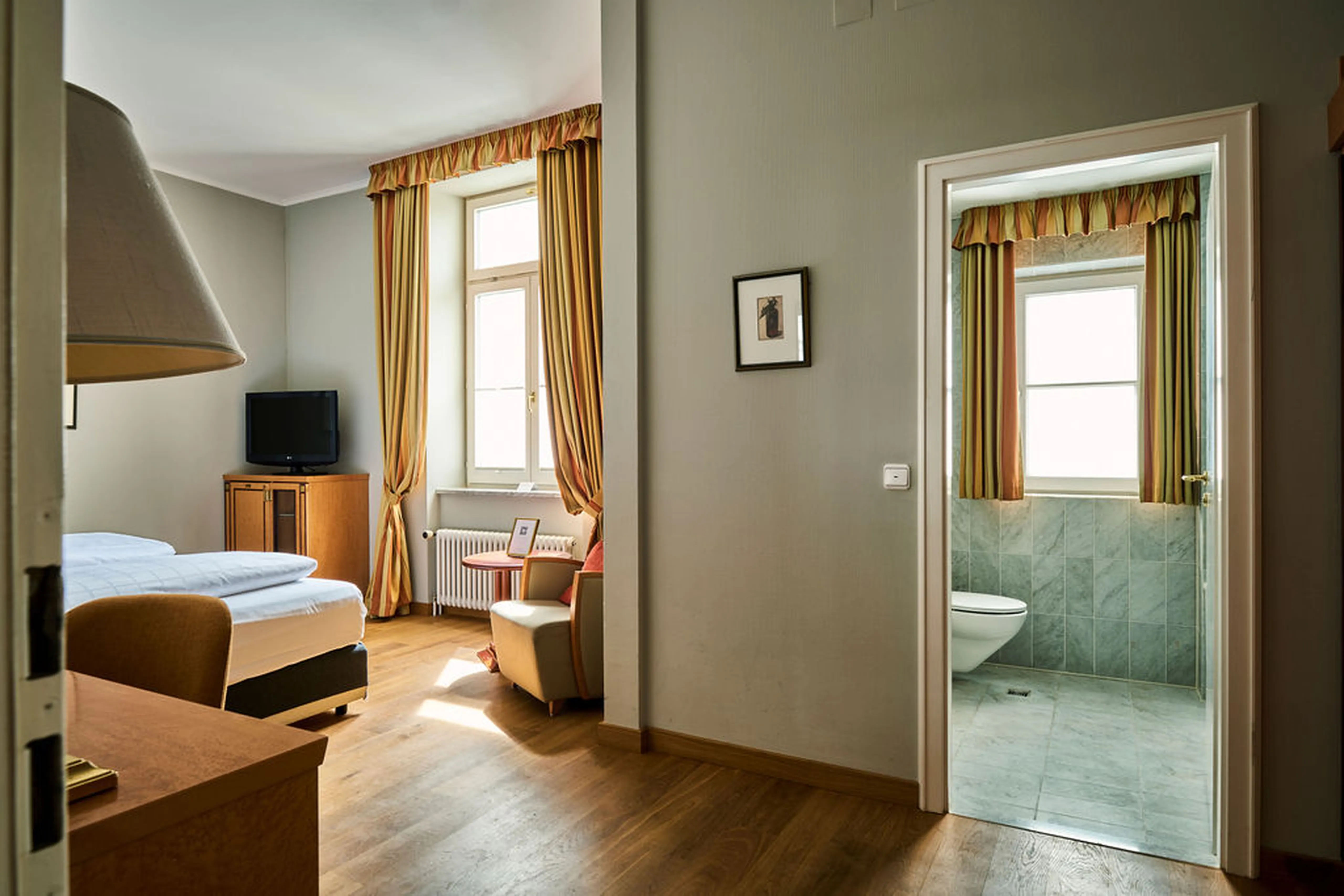 Helles Hotelzimmer mit Holzboden, Doppelbett, Sessel, TV, großen Fenstern mit langen Vorhängen und angrenzendem Badezimmer mit Fliesen.