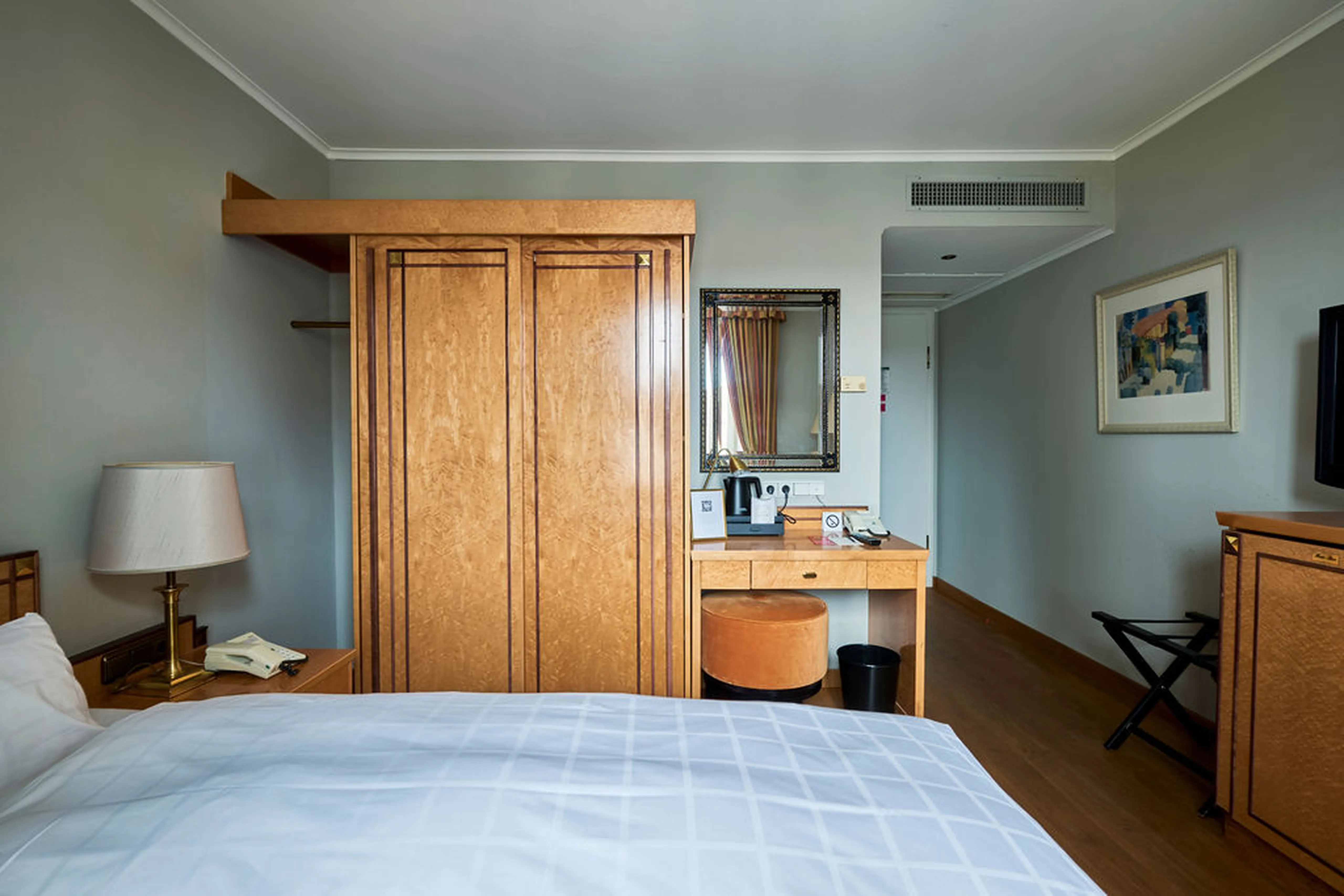 Hotelzimmer mit Holzmöbeln: Kleiderschrank, Schreibtisch mit Spiegel und Hocker, Bett links, Lampe und Kunst an der Wand. Ruhige Atmosphäre.