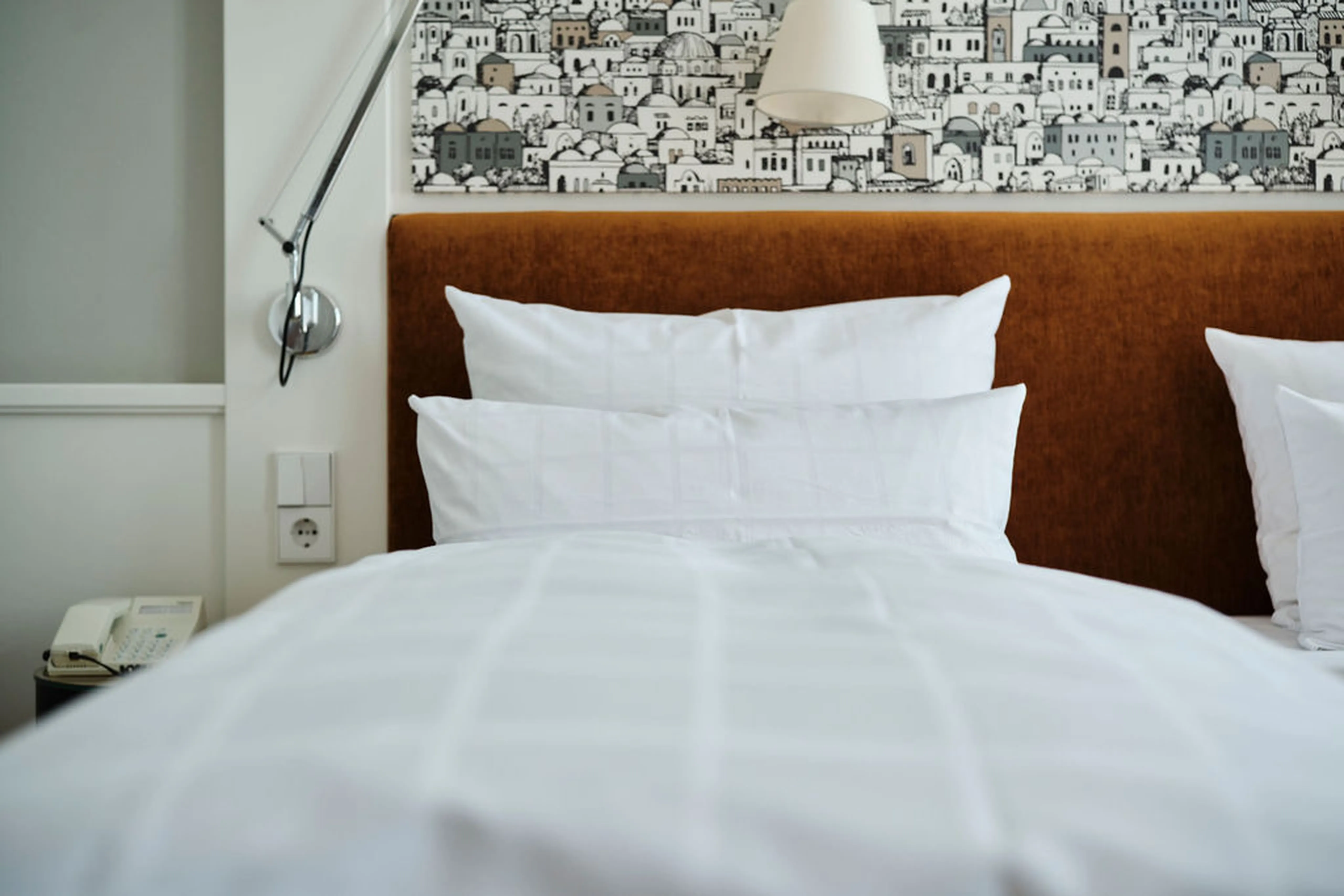 Gemütliches Hotelbett mit weißen Kissen und Bettdecke, einem braunen Kopfteil und einer dekorativen Tapete im Hintergrund.