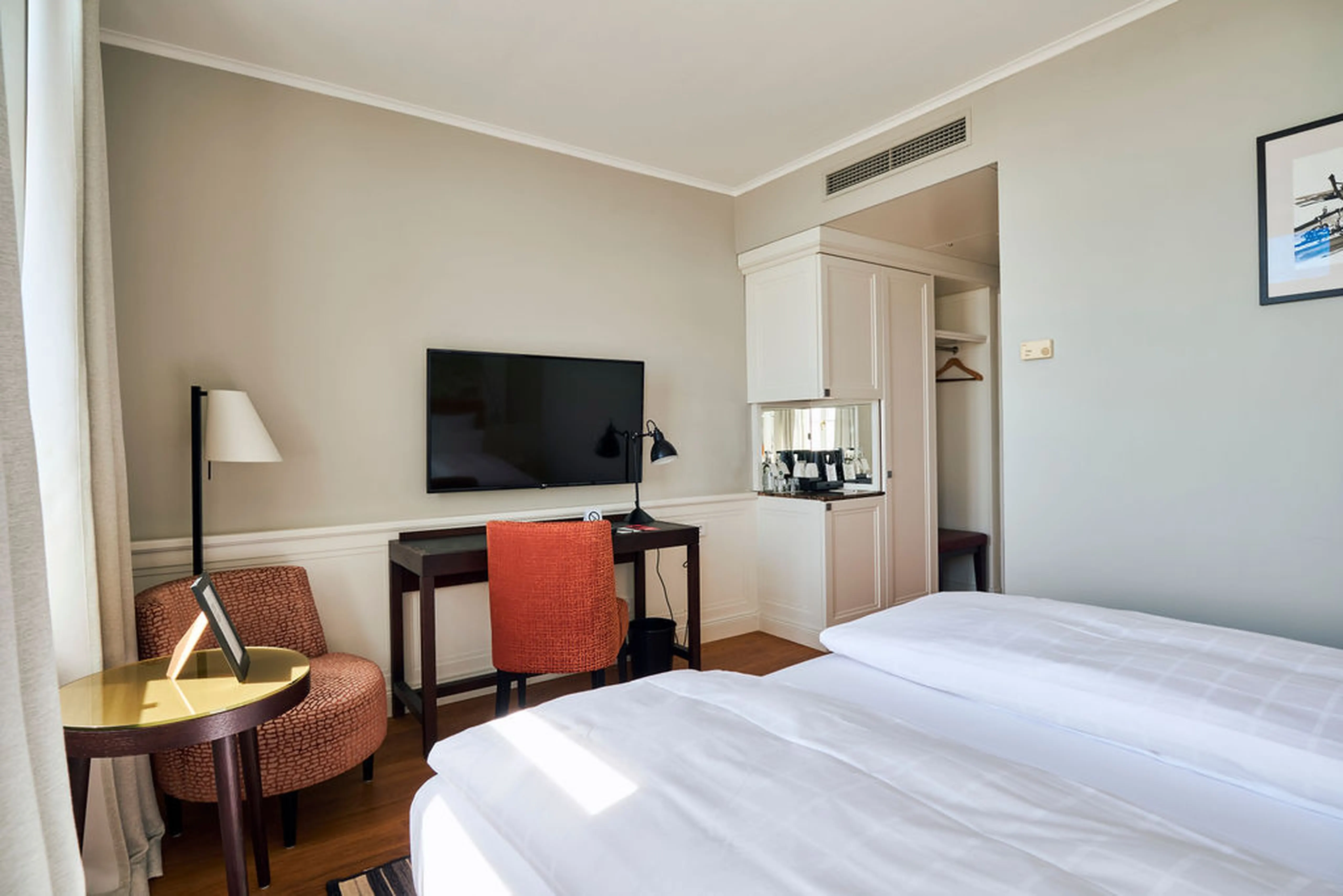 Helles Hotelzimmer mit Doppelbett, Schreibtisch, Flachbildfernseher, rotem Stuhl und einem Spiegel neben der Minibar; gemütlich und modern eingerichtet.