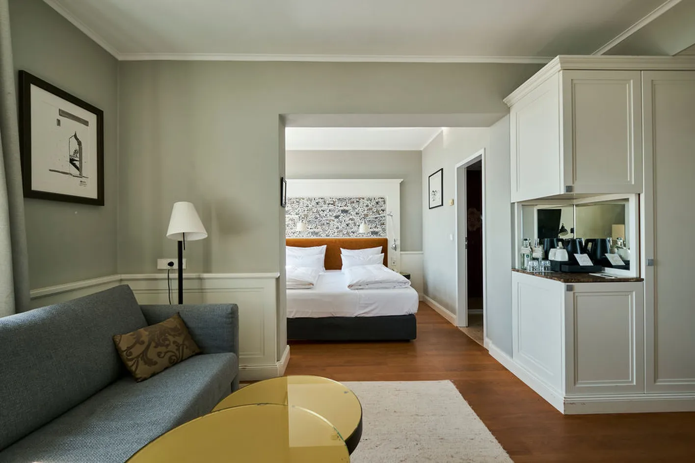 Elegantes Hotelzimmer mit grauem Sofa, gelbem Tisch, Doppelbett und Wandkunst, kombiniert mit schlichten, weißen Schränken und Holzfußboden.