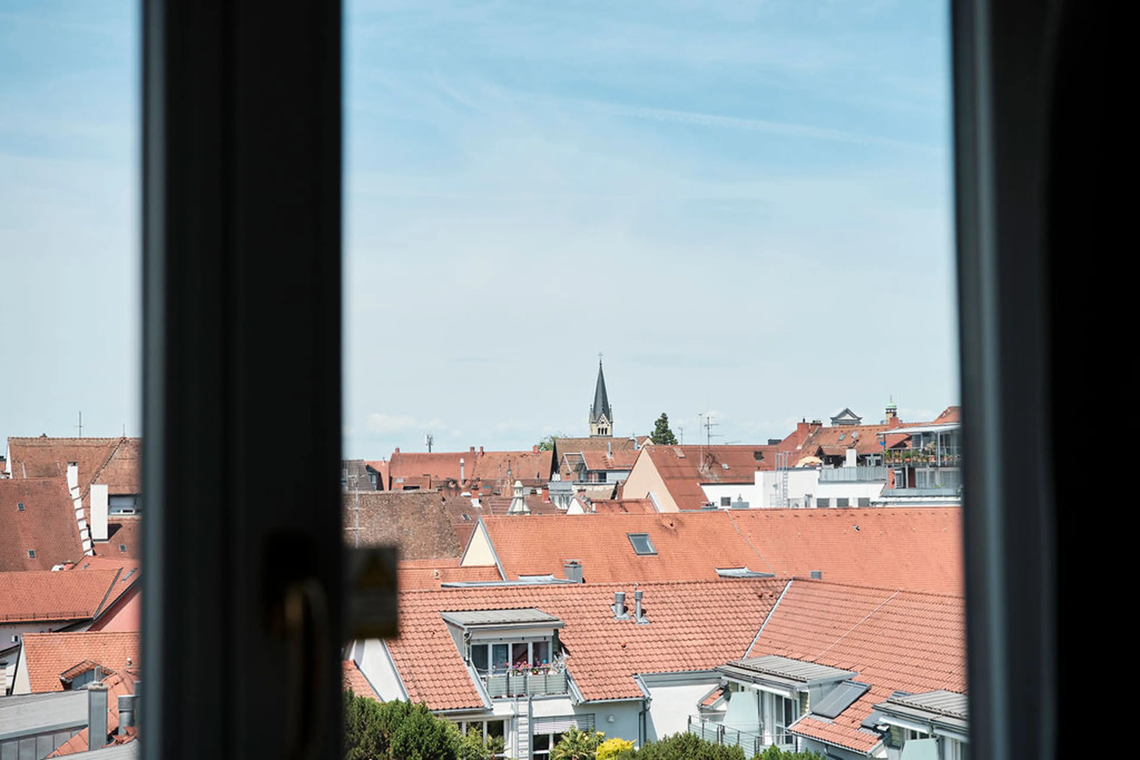 Blick von einem Fenster auf eine Stadtlandschaft mit roten Ziegeldächern und einem Kirchturm bei klarem Himmel.