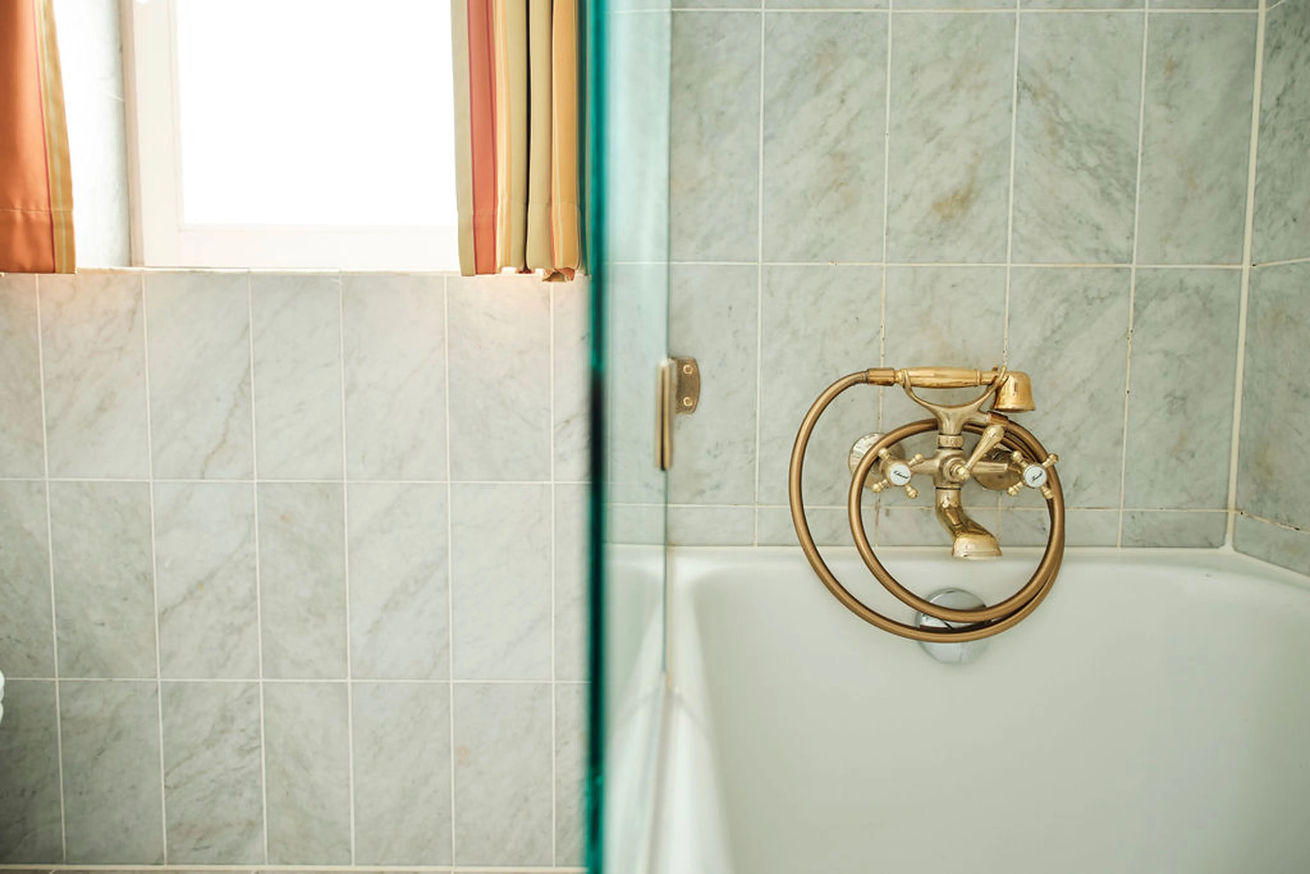 Badezimmer mit Marmorfliesen, einer Badewanne und einem antiken Messingwasserhahn. Vorhang am Fenster links, Glasduschabtrennung sichtbar.