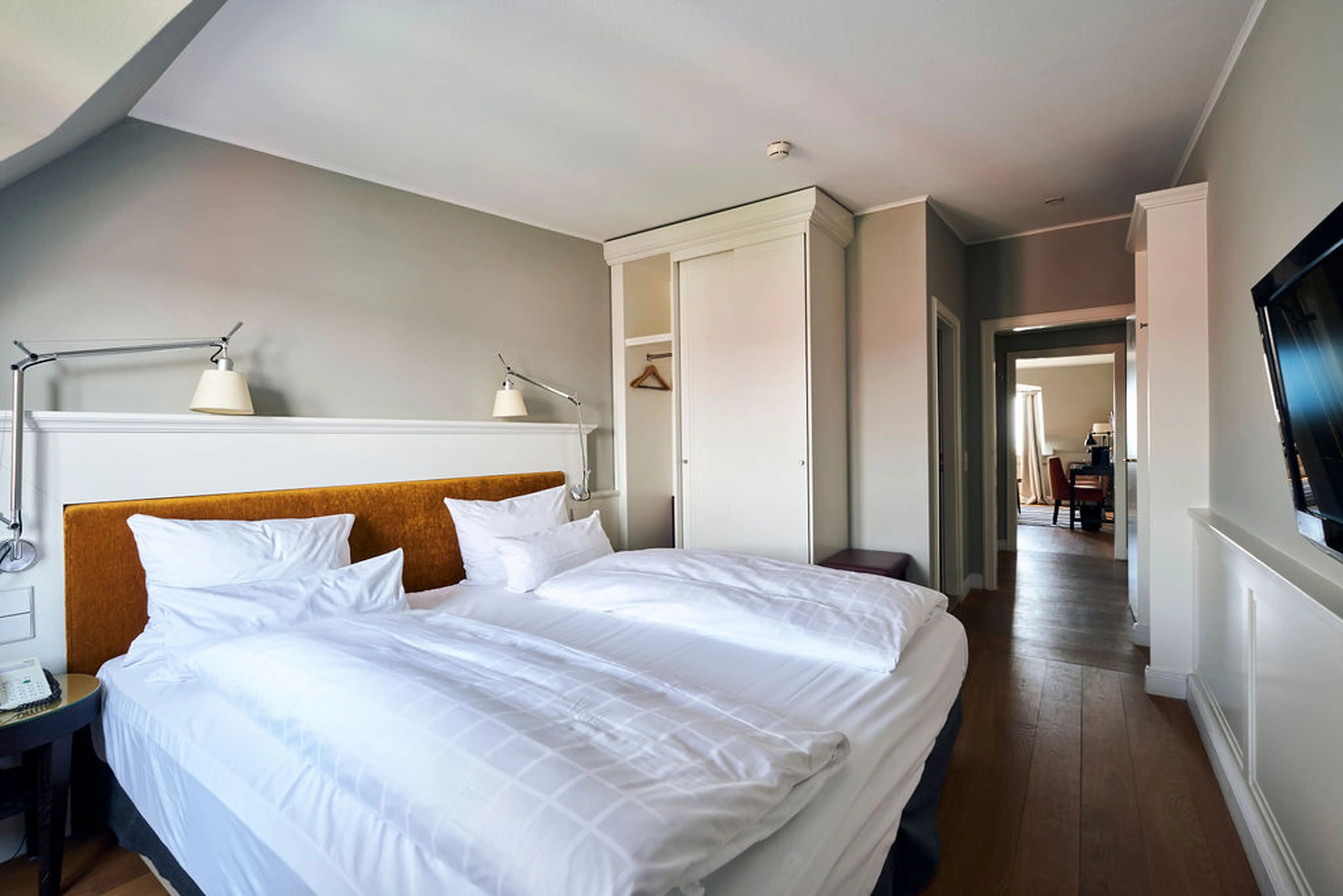 Hell eingerichtetes Hotelzimmer mit Doppelbett, weißer Bettwäsche, zwei Nachttischlampen, einem Schrank und einem Flachbildfernseher an der Wand.