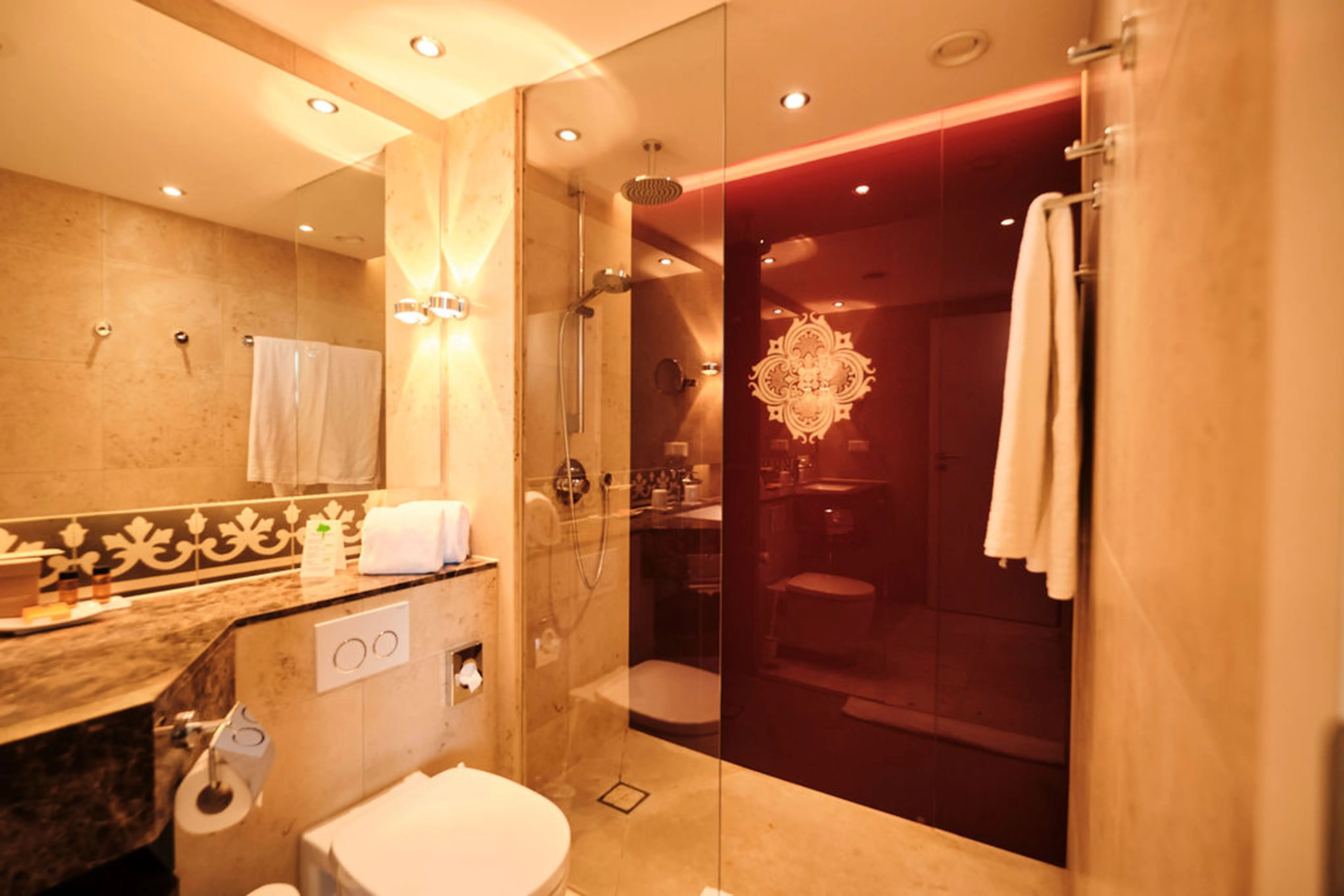 Luxuriöses Badezimmer mit beiger Fliesenoptik, gläserner Dusche, dekorativem Spiegel und Handtüchern; warme Beleuchtung.