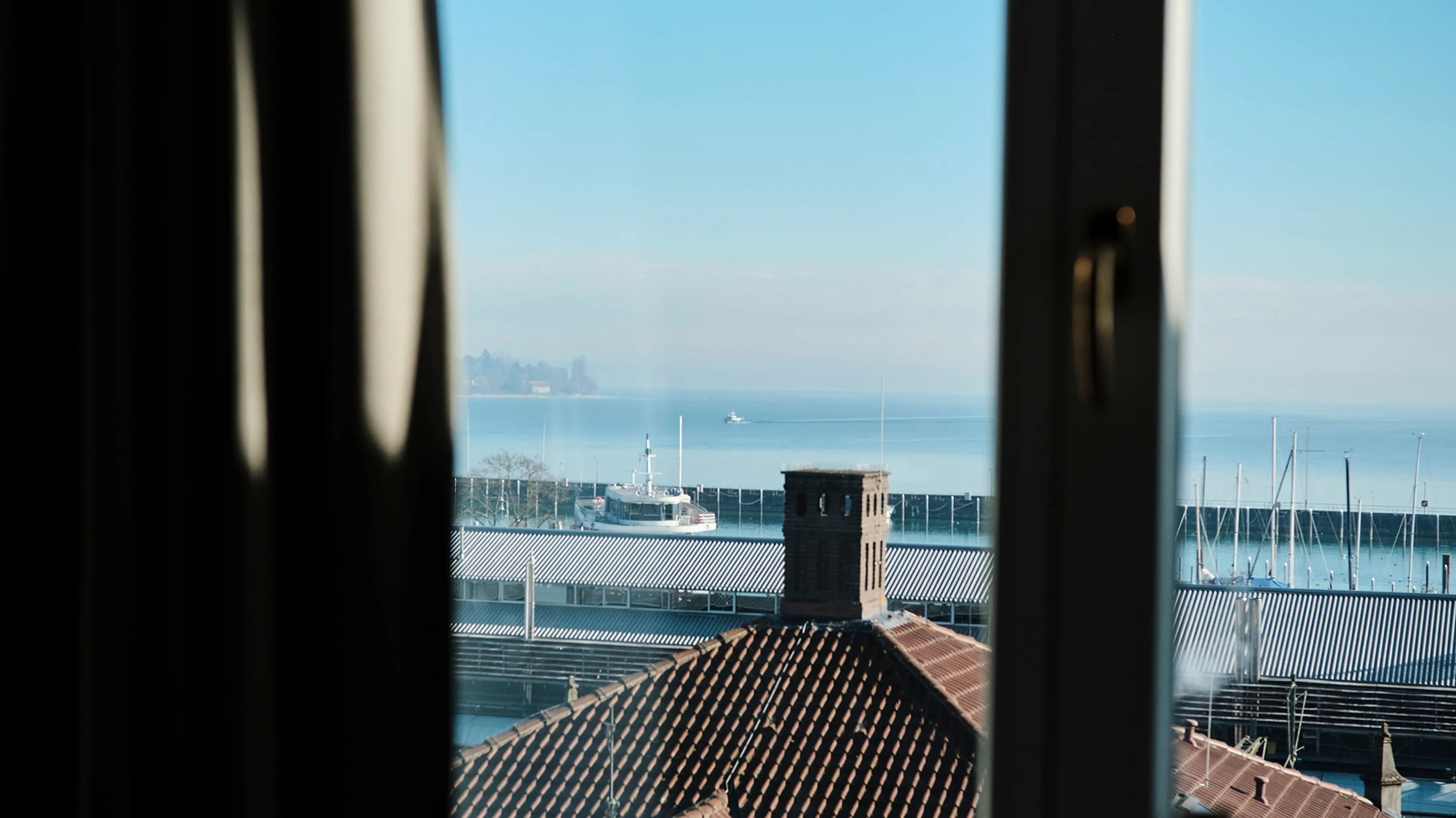 Blick aus einem Fenster auf den Bodensee mit einem Schiff, Dächern und einem Hafen im Hintergrund bei klarem Himmel.
