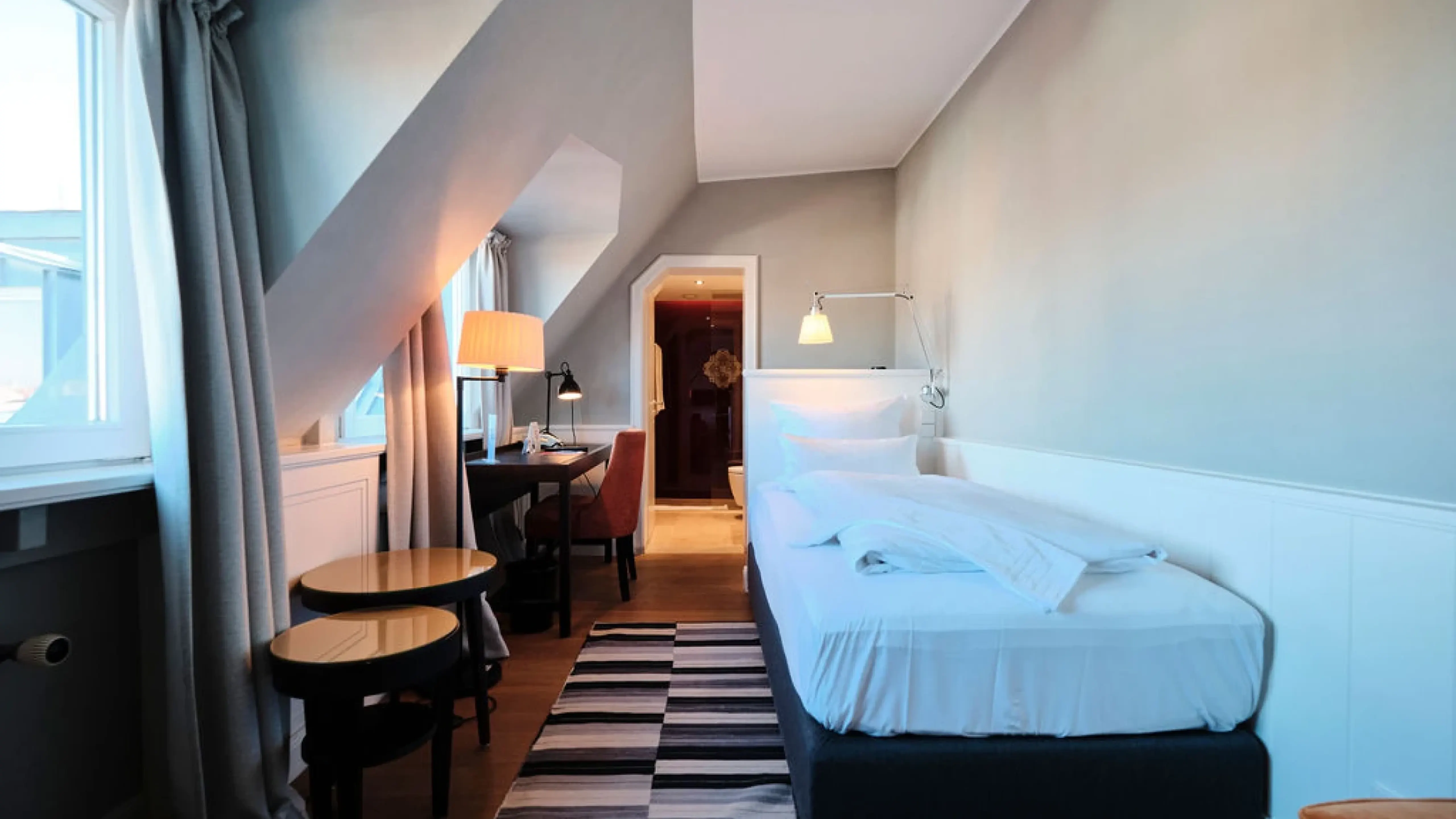Gemütliches Dachzimmer mit Einzelbett, Schreibtisch, Stuhl und Nachttisch. Fenster mit Vorhängen, Teppich und Blick auf Badezimmer.