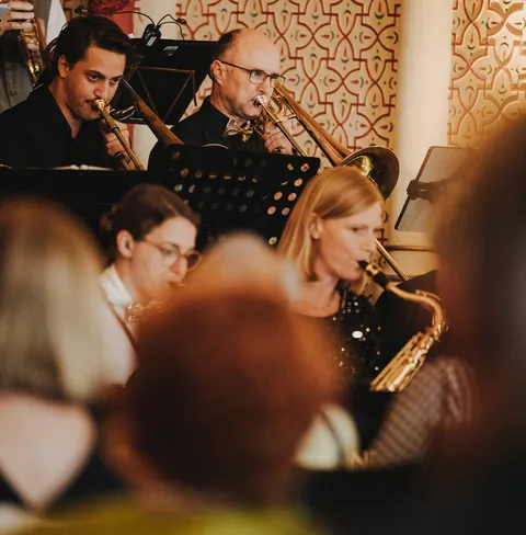 Mehrere Musiker spielen Blasinstrumente in einem Orchester vor einem dekorativen Hintergrund, darunter Posaune und Saxophon.