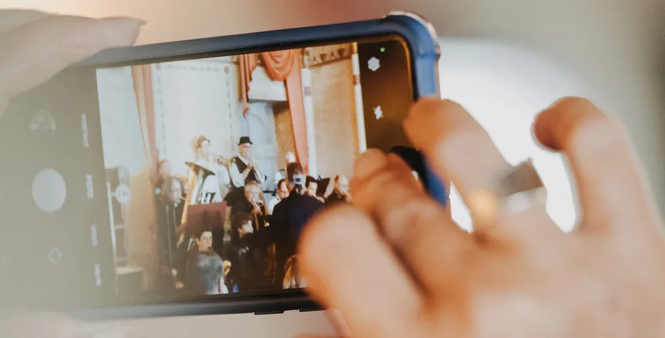 Nahaufnahme von Händen, die ein Smartphone halten, auf dessen Bildschirm ein Orchester bei einem Konzert zu sehen ist.