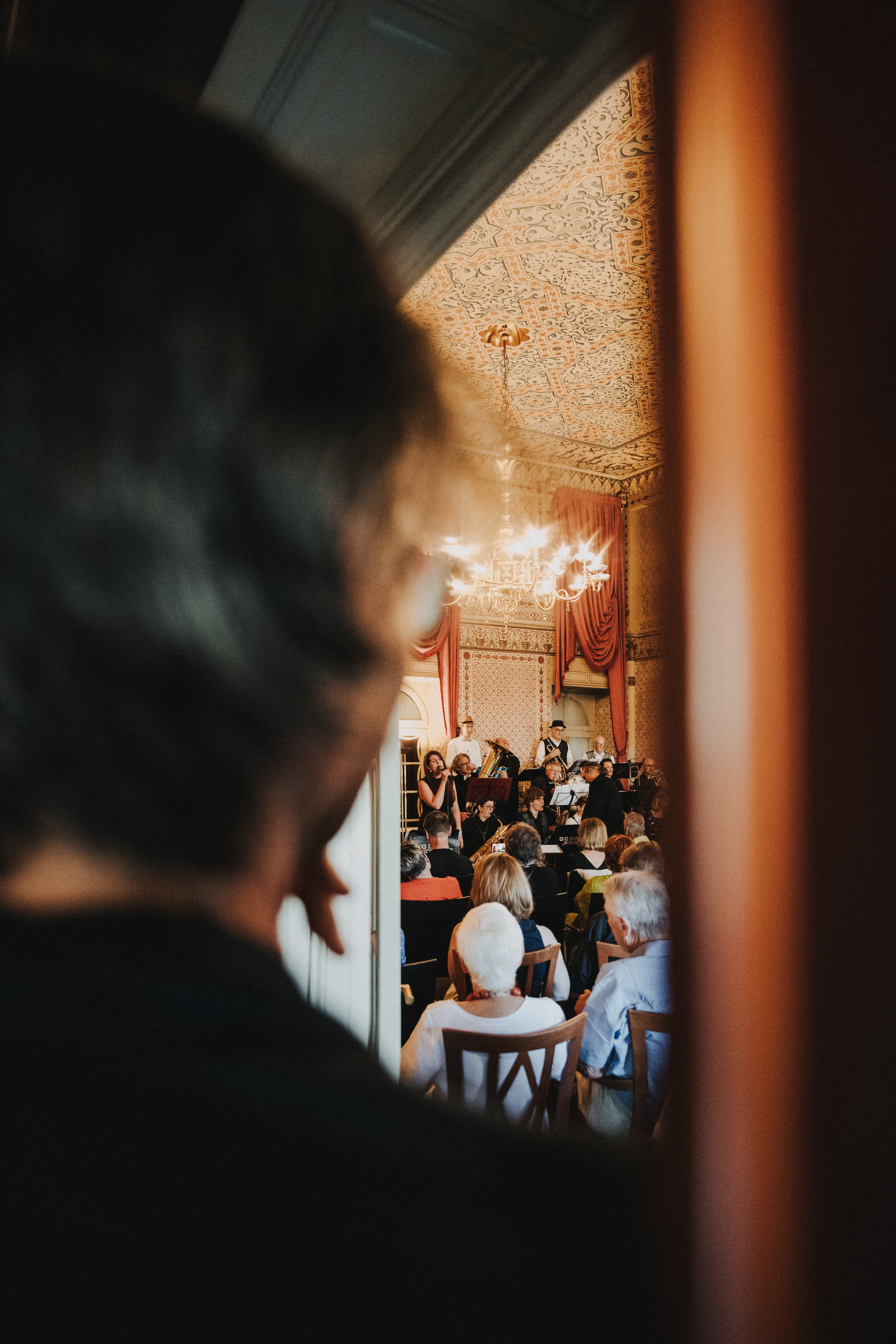 Person schaut durch eine Tür in einen Raum, in dem ein Konzert mit einer Band und Publikum stattfindet. Der Raum ist prächtig dekoriert.