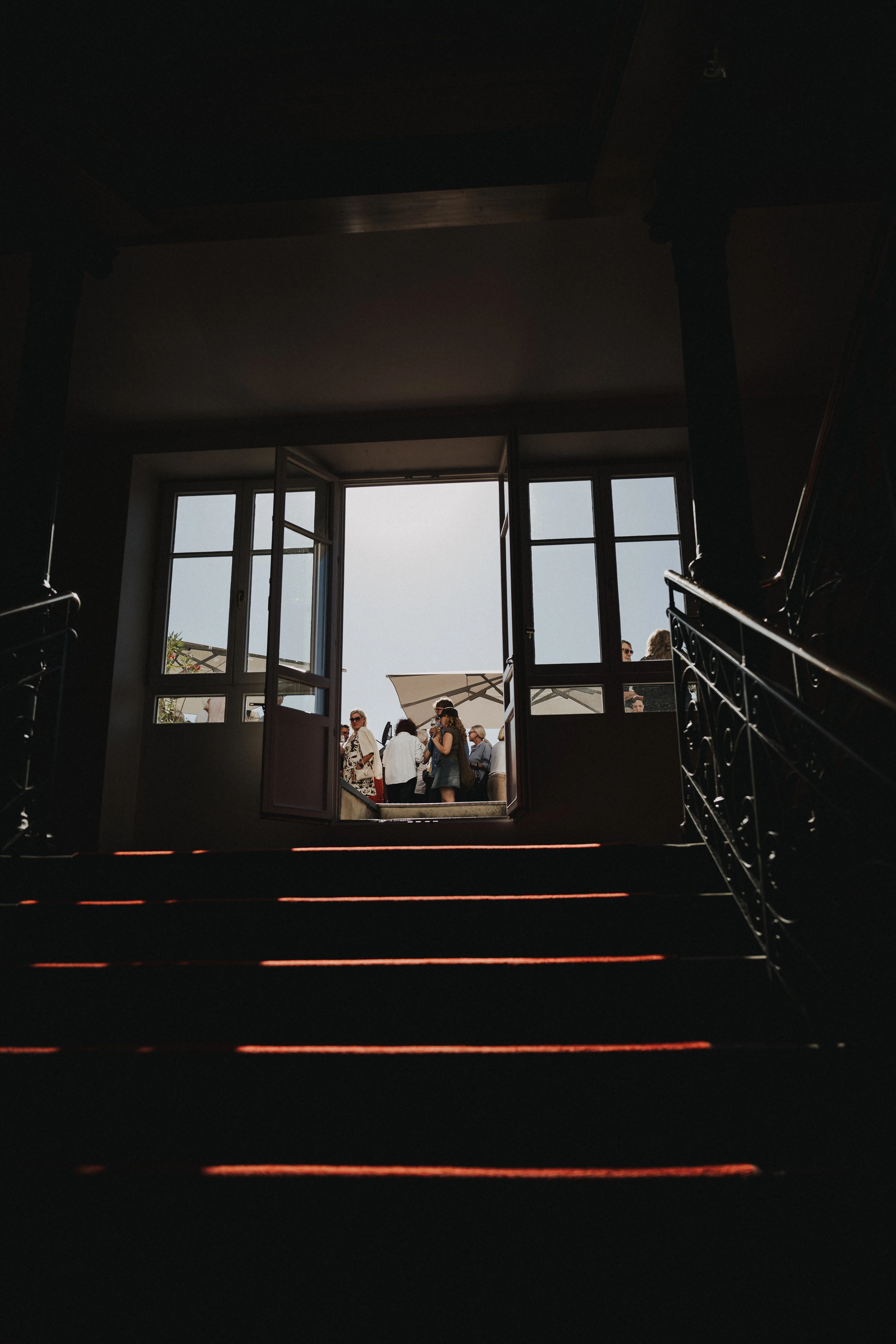 Blick von einer Treppe durch offene Türen nach draußen, wo Menschen bei Sonnenschein unter einem Sonnenschirm stehen.