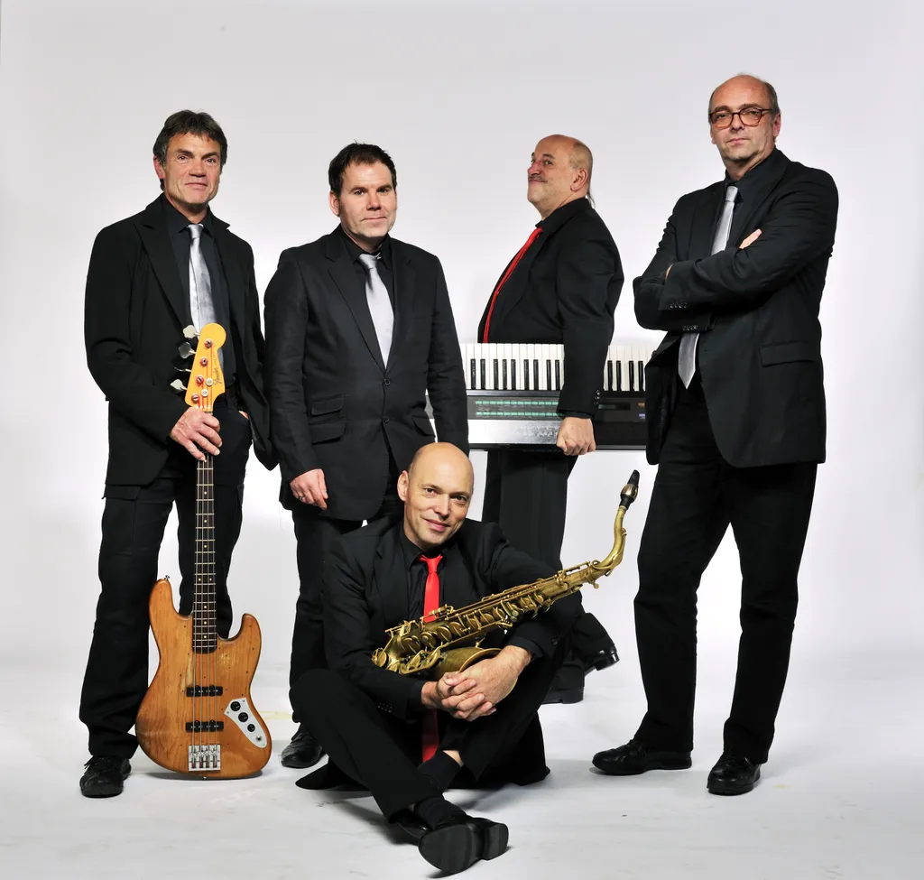 Fünf Männer in Anzügen posieren mit Musikinstrumenten, darunter ein Saxophon, E-Bass und Keyboard, vor einem weißen Hintergrund.