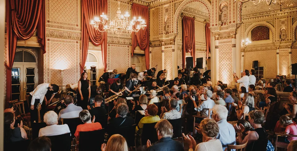 Ein Orchester verbeugt sich vor einem applaudierenden Publikum in einem prunkvollen Saal mit roten Vorhängen und verzierter Decke.