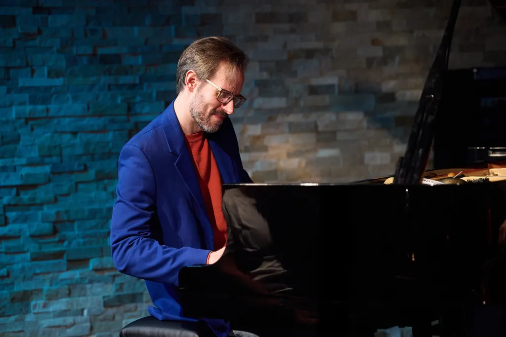 Ein Mann im blauen Sakko spielt lächelnd Klavier in einem Raum mit Ziegelwand und blauer Beleuchtung.
