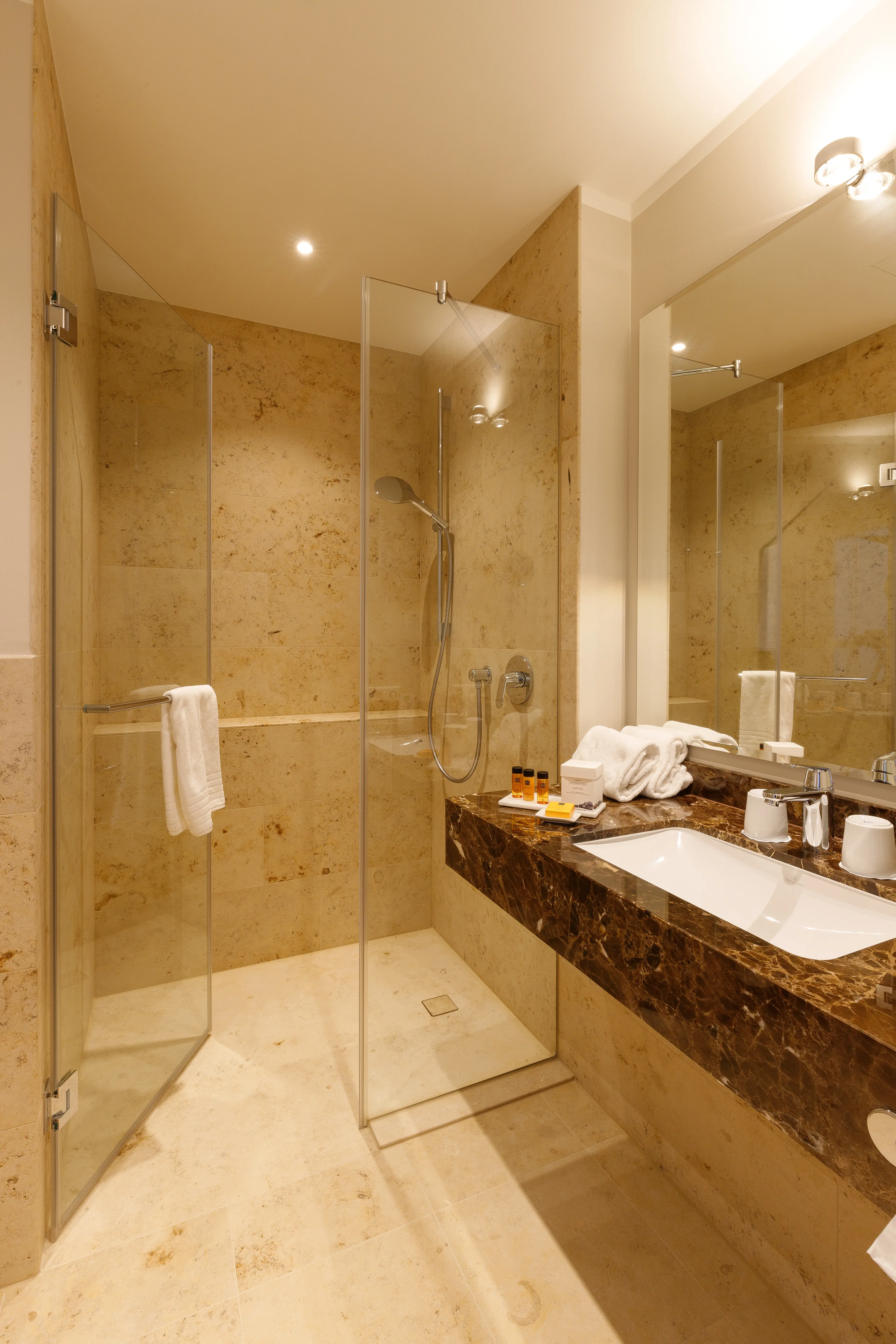 Modernes Badezimmer mit Glassduschkabine, Marmorspüle, Spiegel, Handtüchern und Pflegeprodukten auf einer Ablage. Beige Fliesen an Wand und Boden.